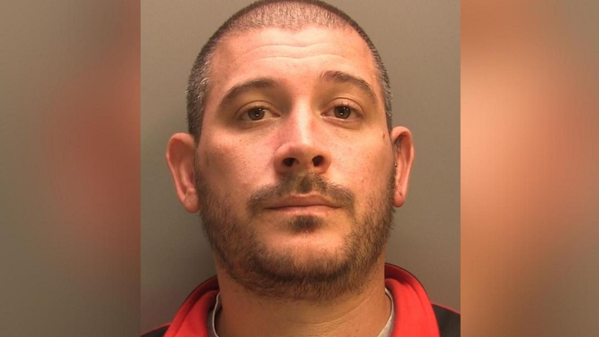 Police custody photograph of James Chater. He has short black hair and beard and is wearing a black and red top and grey T shirt.