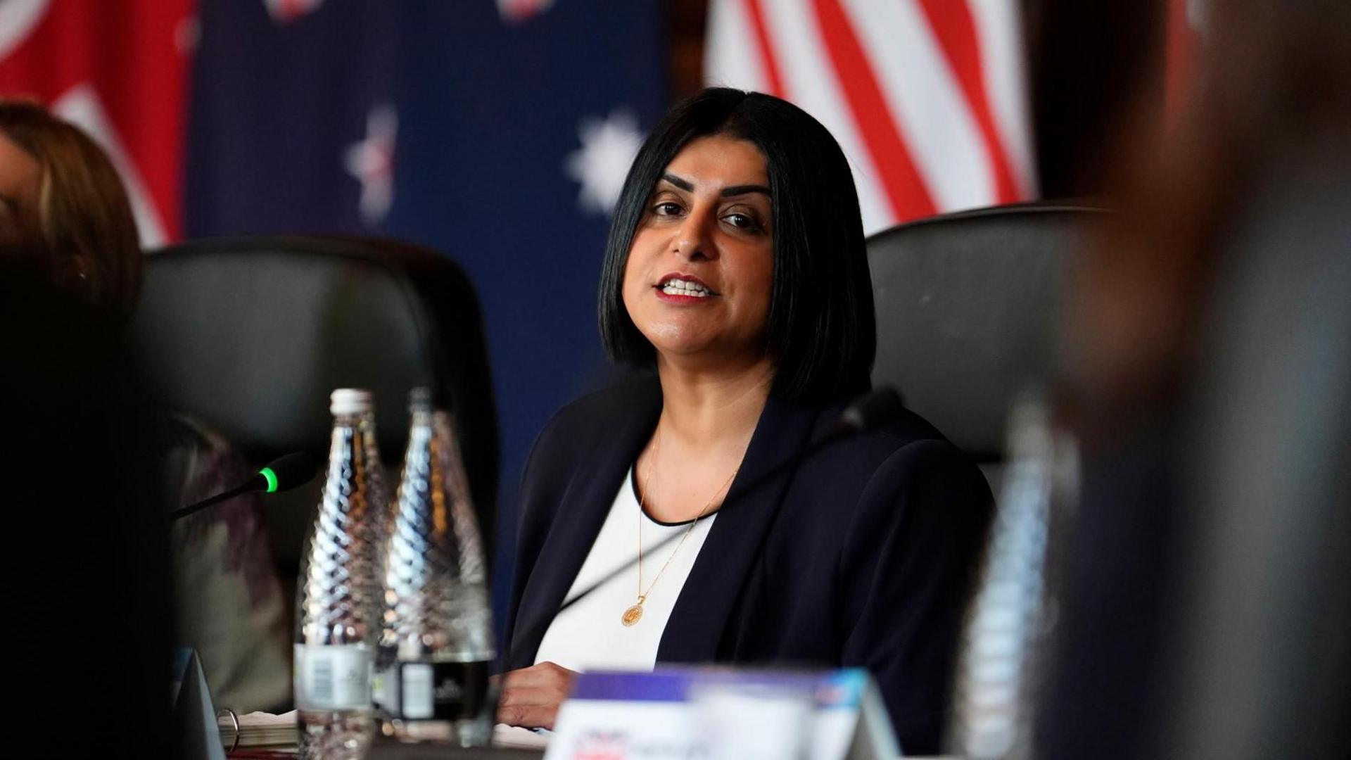 Shabana Mahmood, wearing a dark suit jacket and white top, sits at a meeting room table. The flags of the US, Australia, New Zealand and the UK are draped in the background.