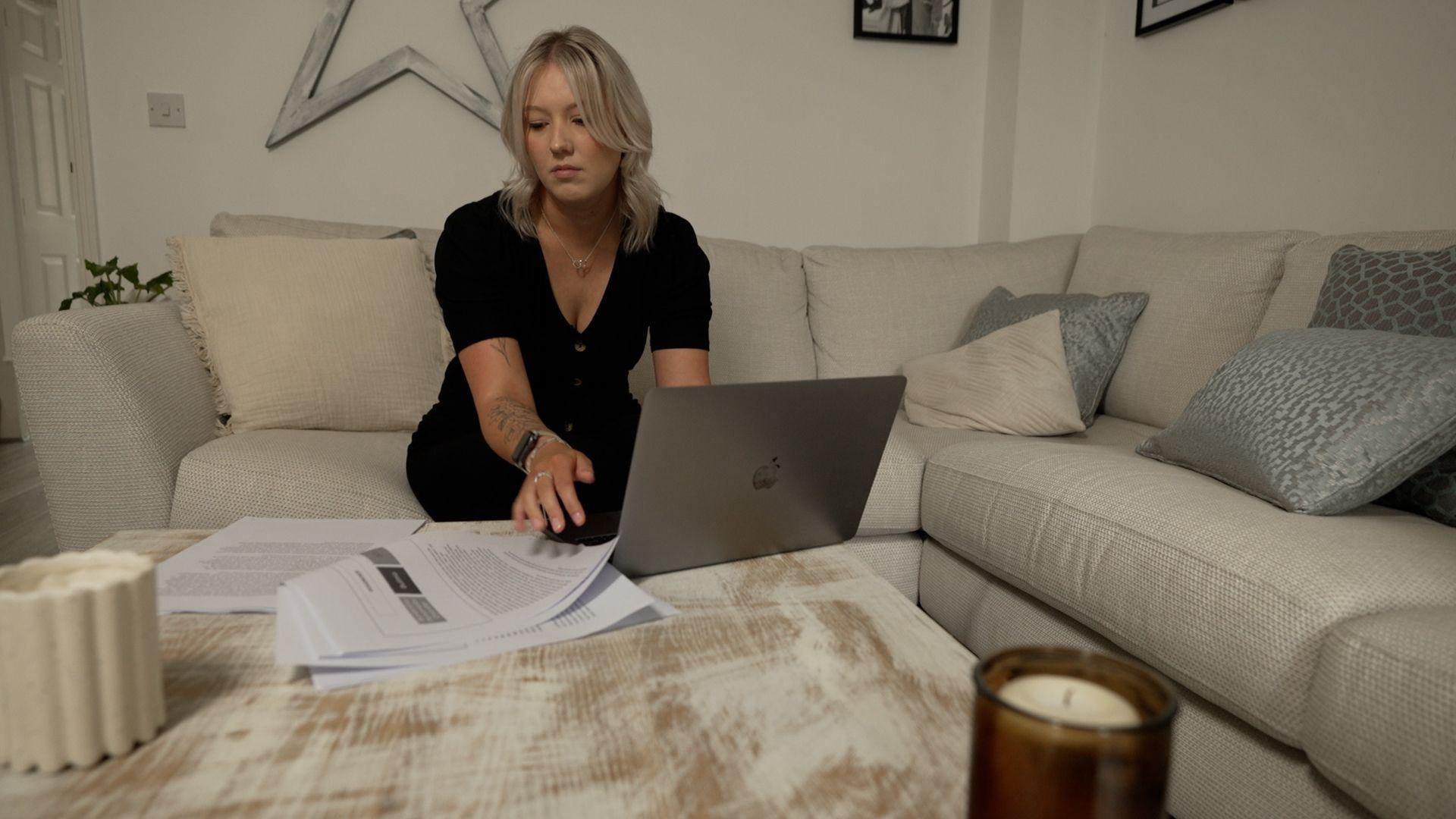 Jessica Matthews looks at a stack of paperwork related to her diagnosis. It sits next to her laptop. She has a worried expression on her face and is in her living room.