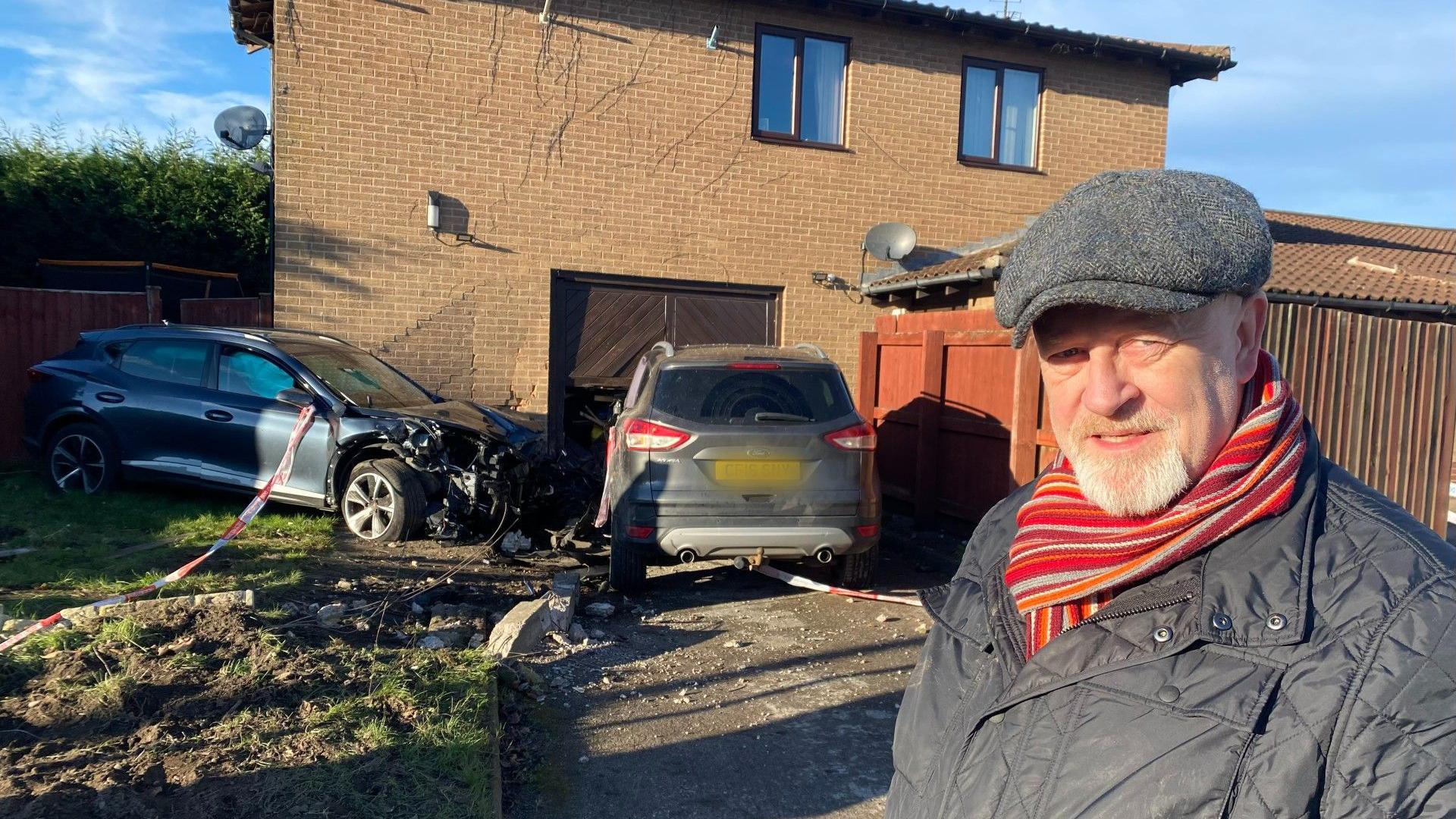 A man looks at the camera in front of two damaged cars outside Calverton Miners Welfare. He is wearing a red and orange striped scarf tucked in to a black padded coat. He is also wearing a grey hat.