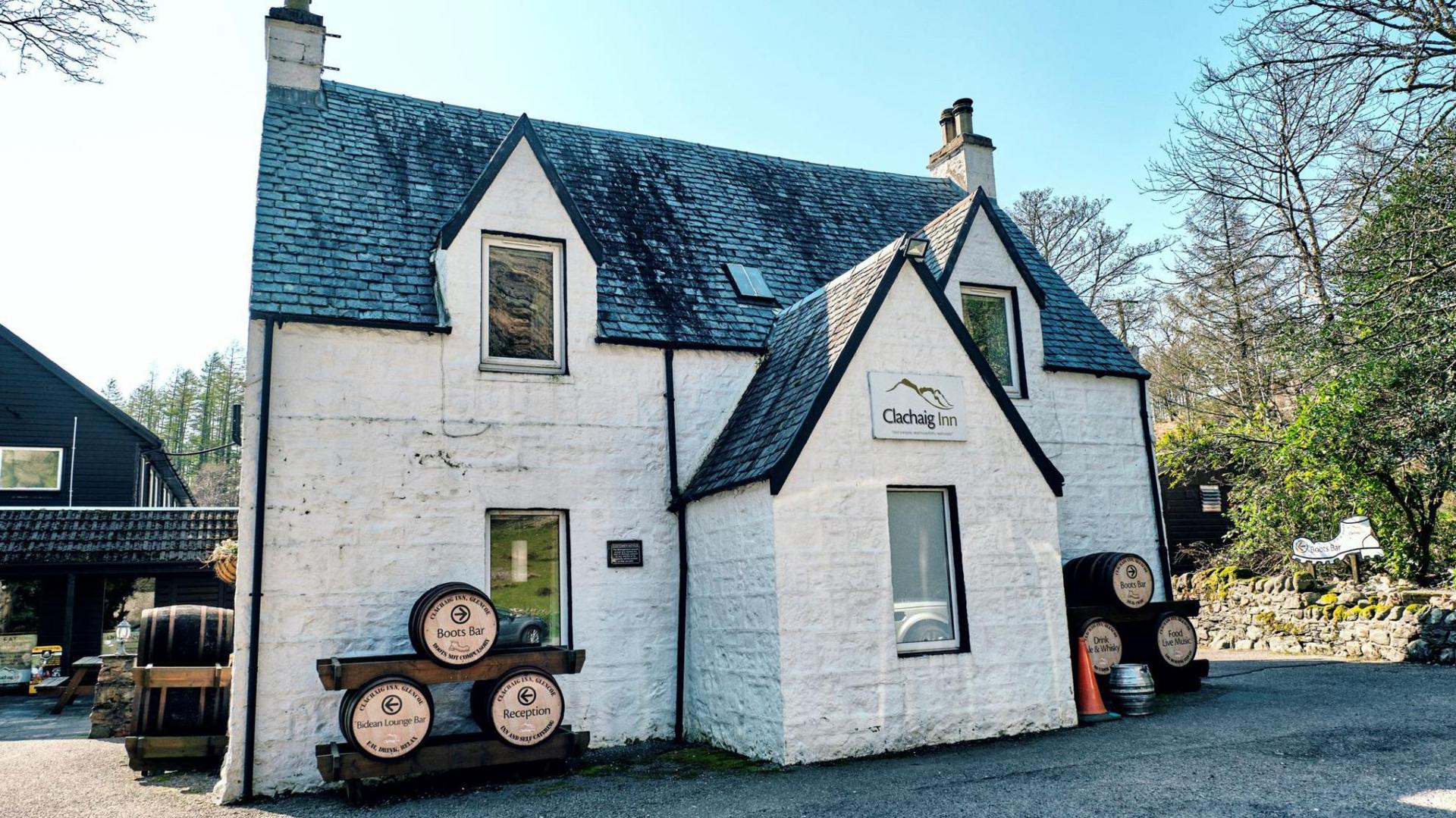 The inn is a white-walled, slate roof building. There is a sign above a narrow window that reads: "Clachaig Inn" and has a silhouette of a mountain. There are three old beer barrels advertising the Boots Bar.