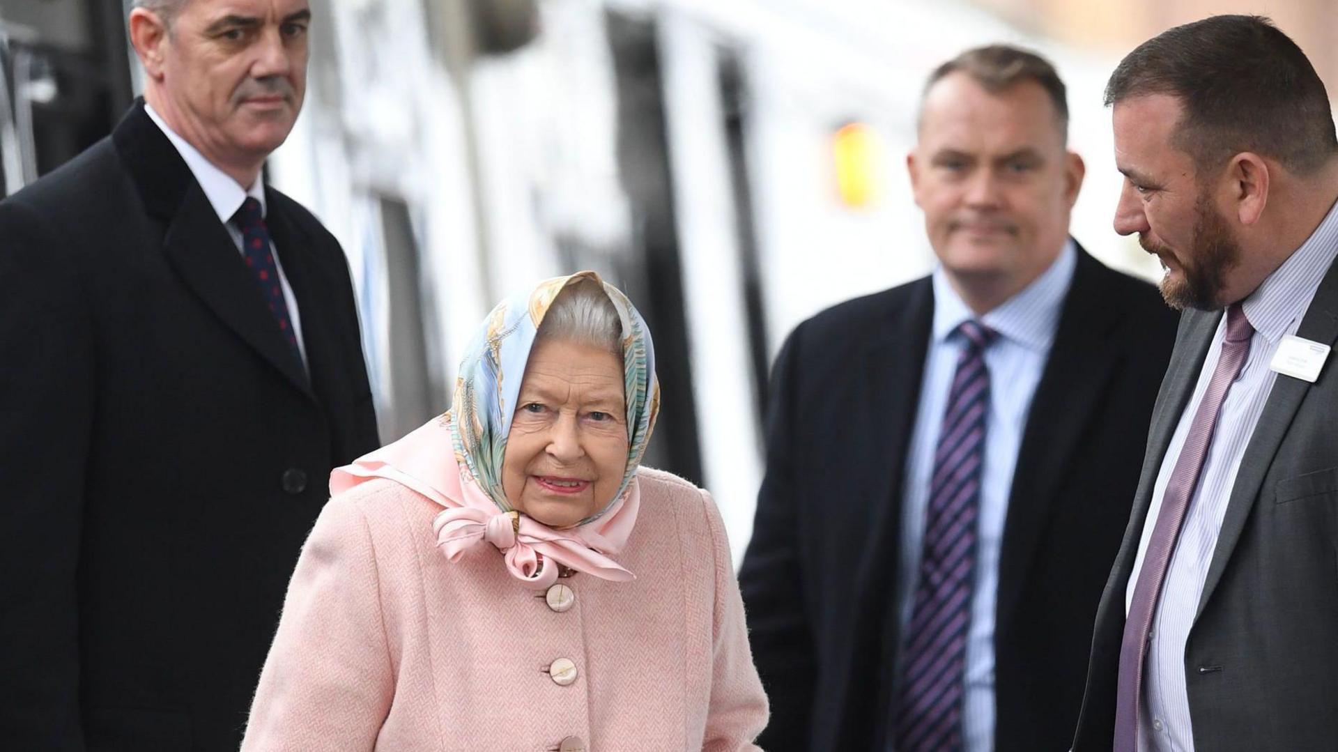 Queen Elizabeth II stands on a station platform surrounded by three men in suits. She wears a pale peach coat with matching peach and pale blue headscarf.