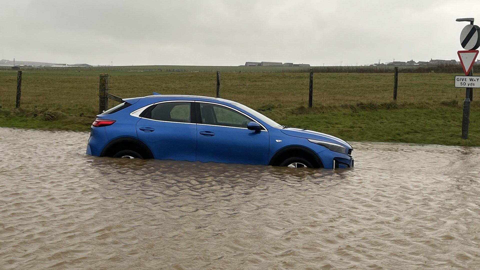 A blue car sitting in a big pool of water. Green fields in the background.