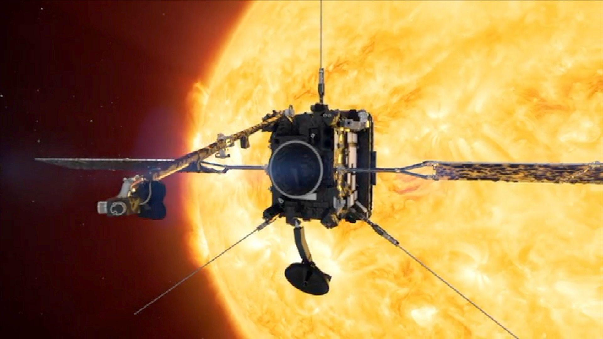 The spacecraft appears as a grey box with an antennae sticking upwards and pointed diagonally outwards from the bottom corners and two solar panels protruding from each side. Behind it appears a raging yellow Sun