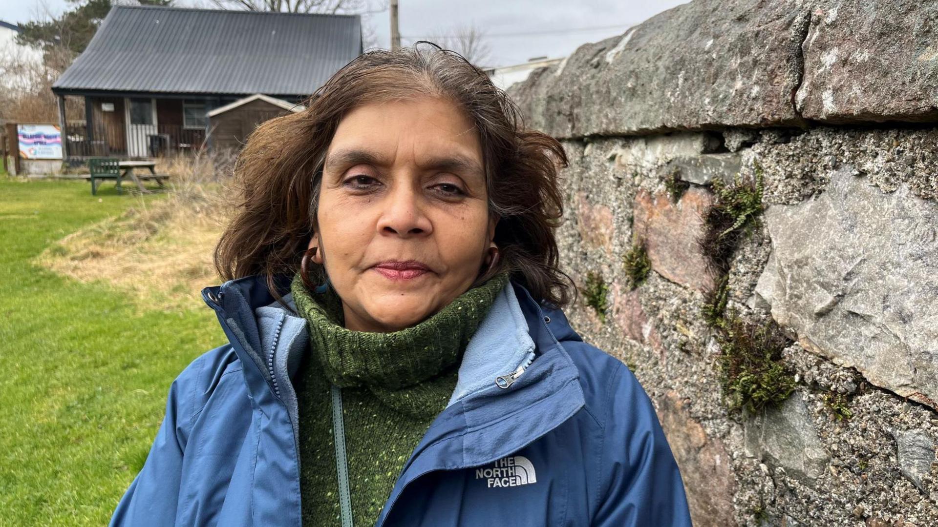 Mamta Patel is wearing a green turtle neck jumper and a blue rain jacket. She is standing next to a stone wall. There is a building with a corrugated roof in the background.