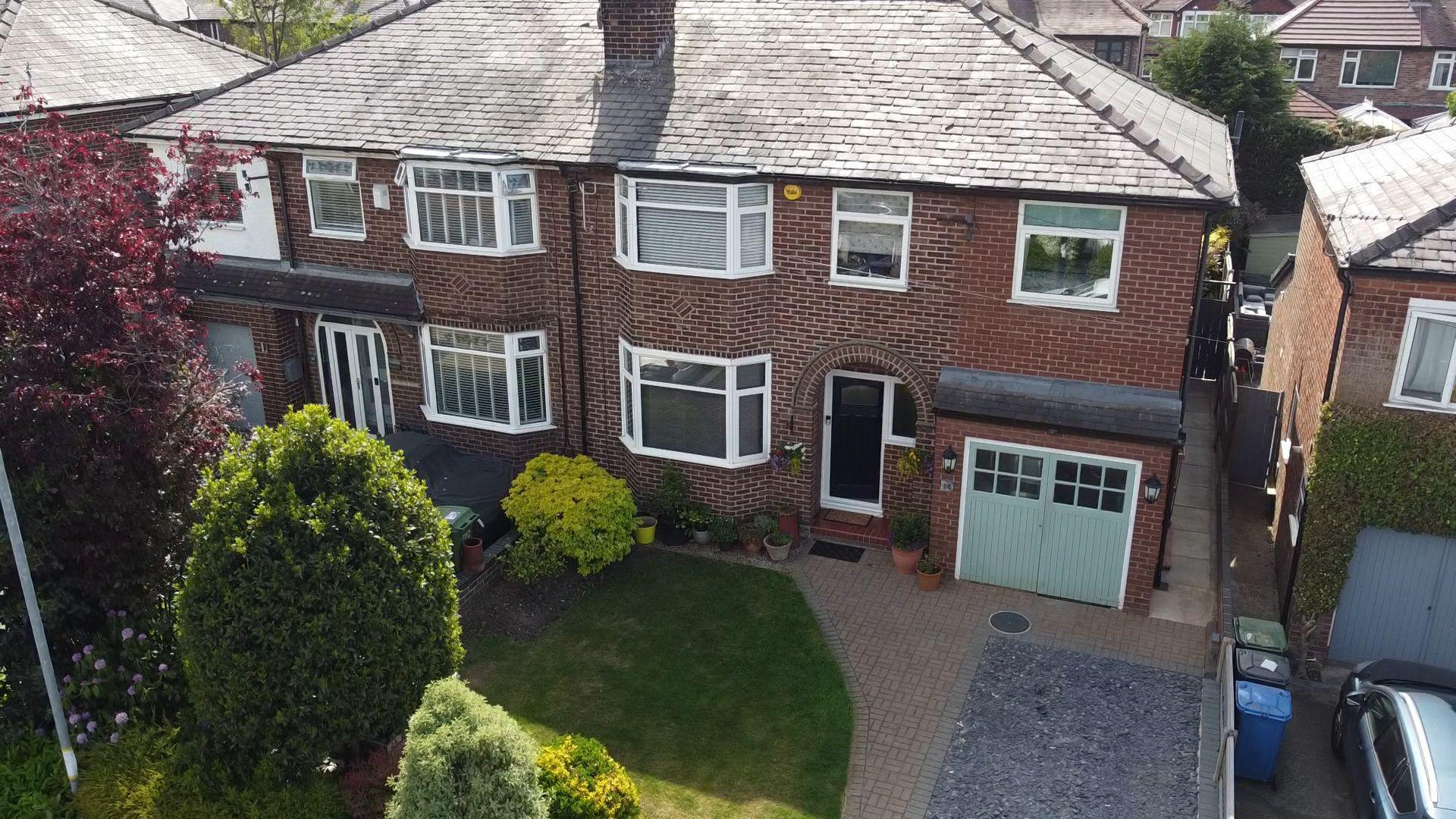 A brown brick semi-detached house with a light green garage door and a front garden with a lawn, trees and bushes.