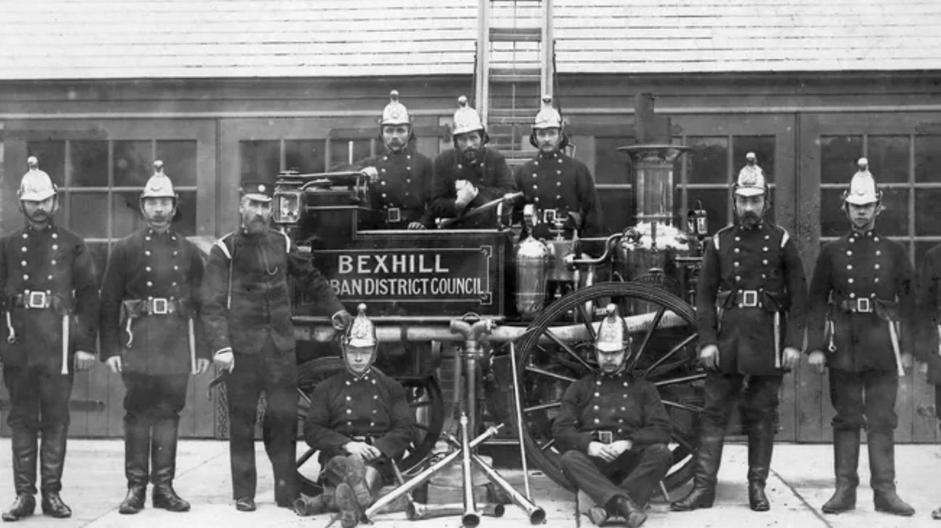 Black and white image of eleven men lined up with the small fire engine. They are dressed in uniform, with long boots and long jackets with 12 buttons, and a wide belt with a large buckle. They are also wearing tall helmets that curve slightly at the top