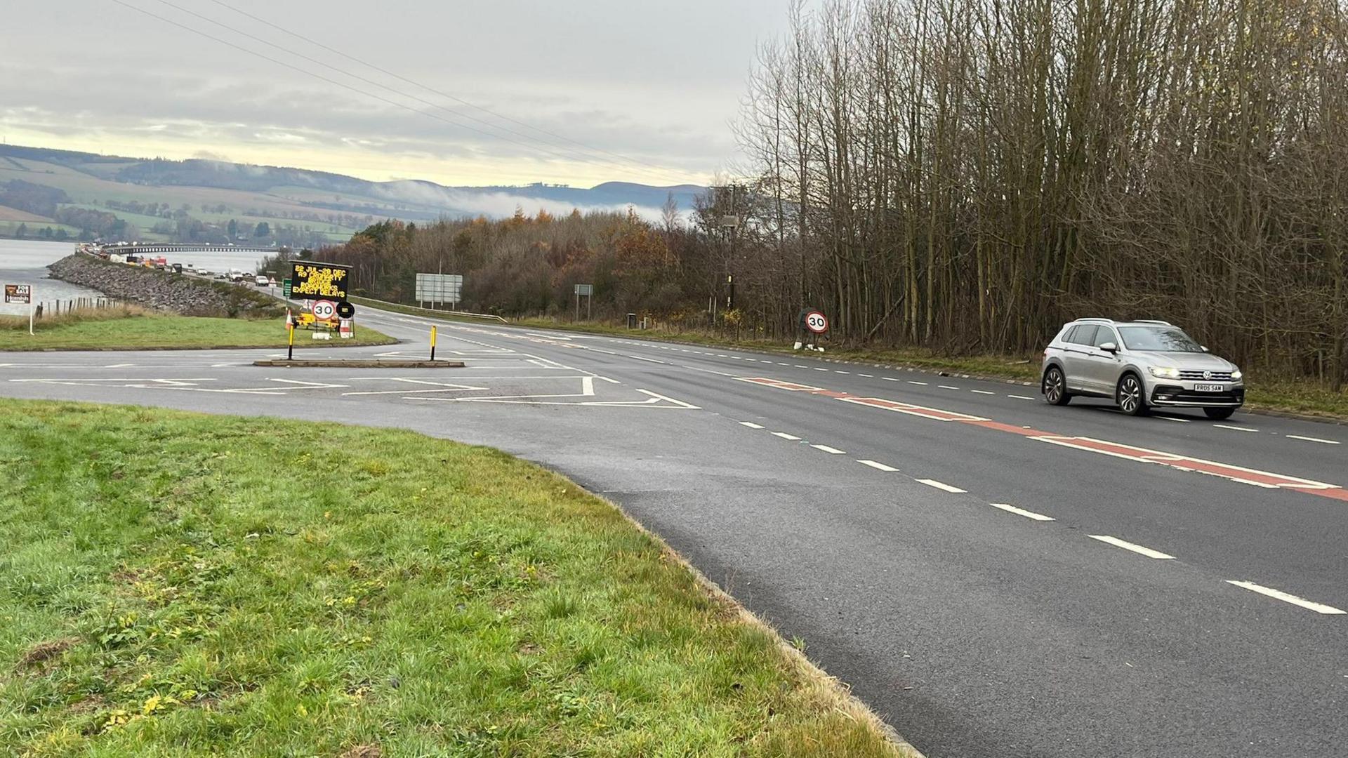 A junction on the A9 with the Cromarty road bridge and Cromarty Firth in the distance. A silver SUV is passing the junction.