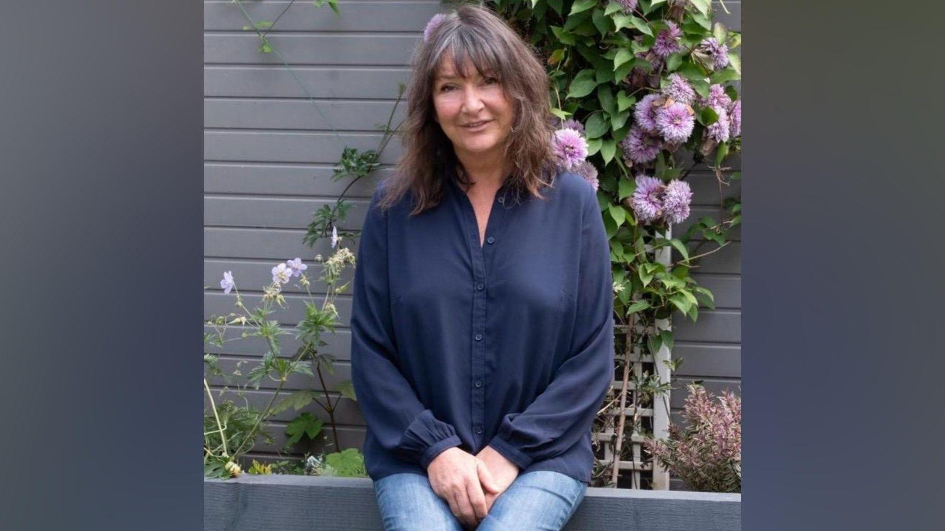 A woman in a blue shirt and jeans sits on a garden wall with flowers behind her