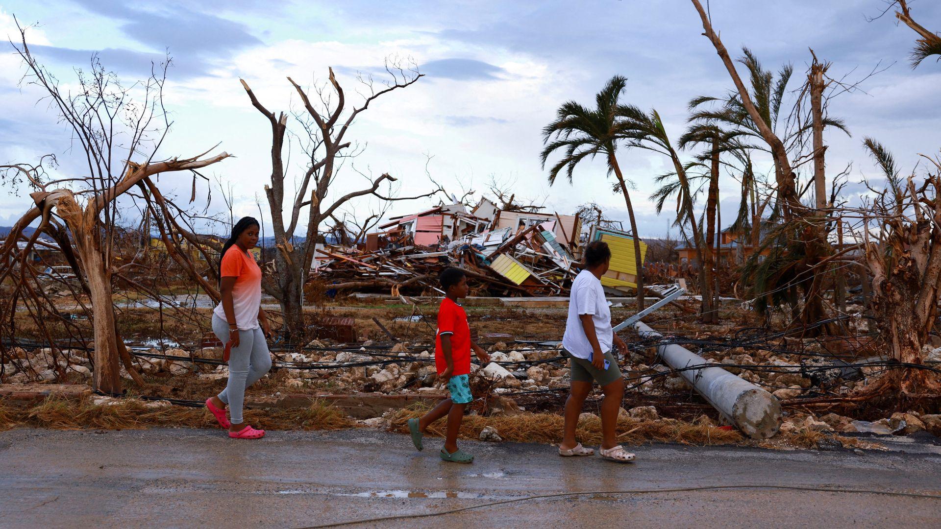 Two women and a boy walk along a road. Behind them, building rubble and electricity poles are laid on the ground. The trees are badly damaged with just a few palm trees left standing. One of the two women is wearing a t-shirt which fades from orange to white, has long black hair, grey jogging bottoms and pink crocs. The boy walking ahead of her is wearing a red t-shirt, aqua shorts and green crocs. The woman in front of him is wearing a white t-shirt, green shorts and cream crocs.