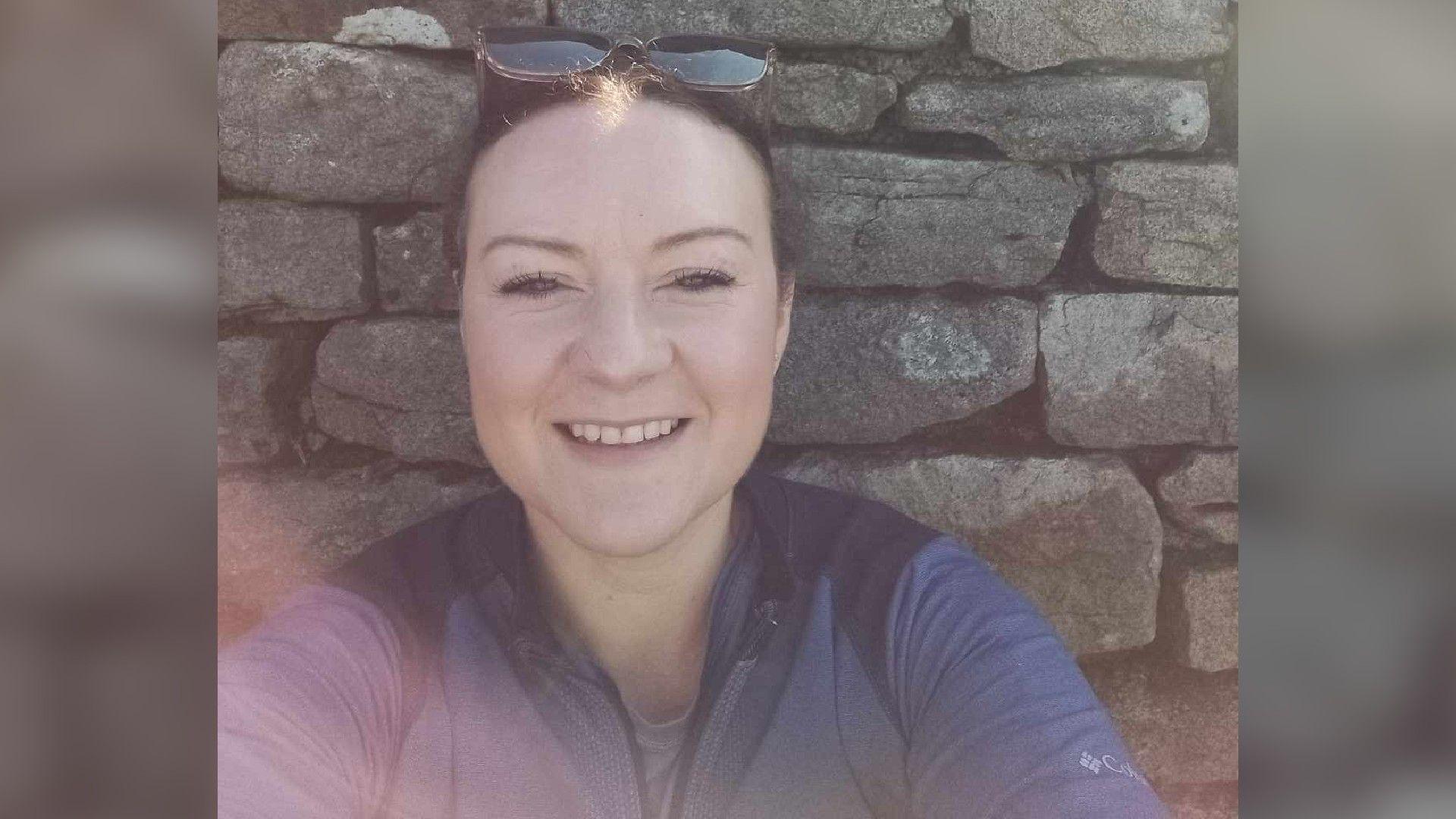 A woman in a dark coloured top with sunglass on top of her head smiles at the camera as she sits in front of a dry stone wall