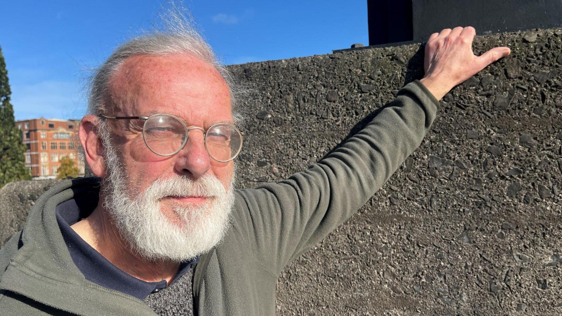 John Anderson is standing with his hand on a wall. He has a white beard and is wearing glasses and a grey hooded top.