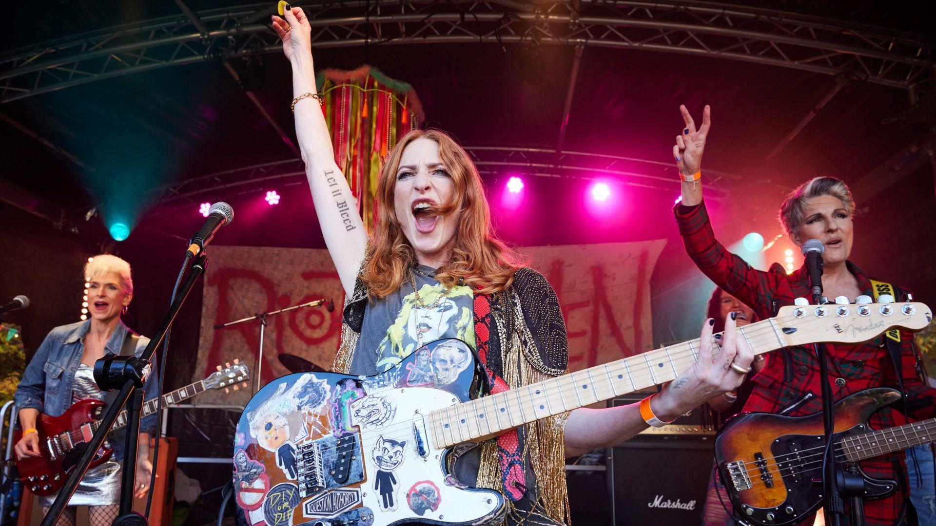 From left to right Amelia Bullmore, Rosalie Craig and Tamsin Greig. Amelia is holding a red Gibson guitar. She has bleach blonde hair and is wearing a denim jacket over a shiny silver dress. Rosalie has a Fender guitar covered in stickers. She is wearing a black T-shirt with an image of Debbie Harry on the front. She has her right arm raised, revealing a tattoo reading Let It Bleed. Tamsin is wearing a red tartan jacket and holding a sunburst bass guitar. She has her right arm raised and is making a peace symbol with her fingers.