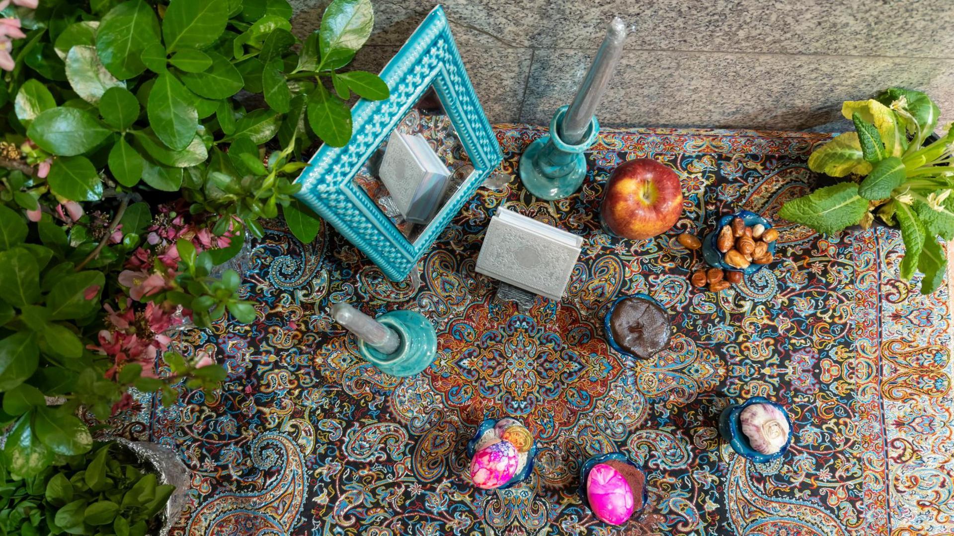 Aerial shot of 7 items arranges on an elaborately patterned rug, with candles and a mirror. Identifiable are an apple, nuts, garlic and a painted egg. There are green plants placed to the side of the display.