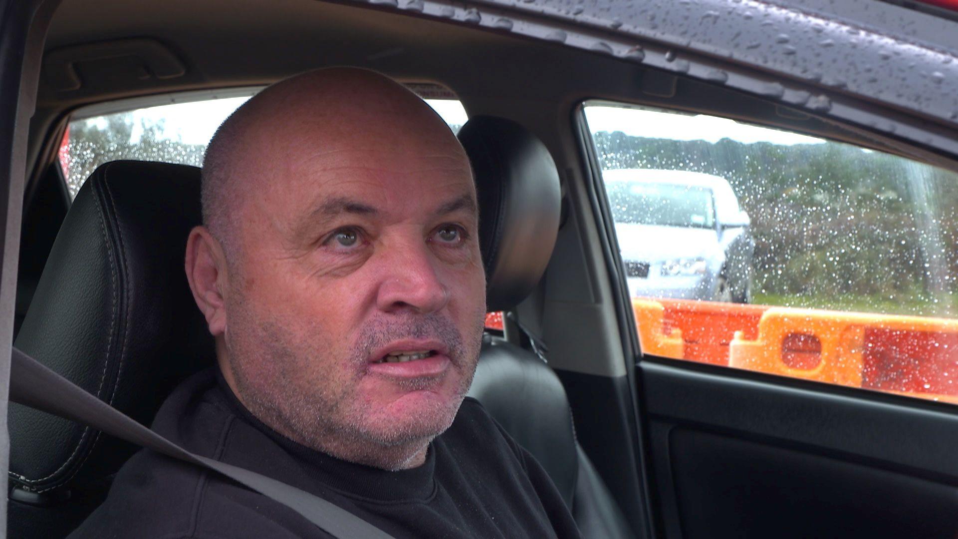 Tommy Hart inside his car wearing a black t-shirt and a seatbelt. Through the passenger window is orange and red barriers on the road. 