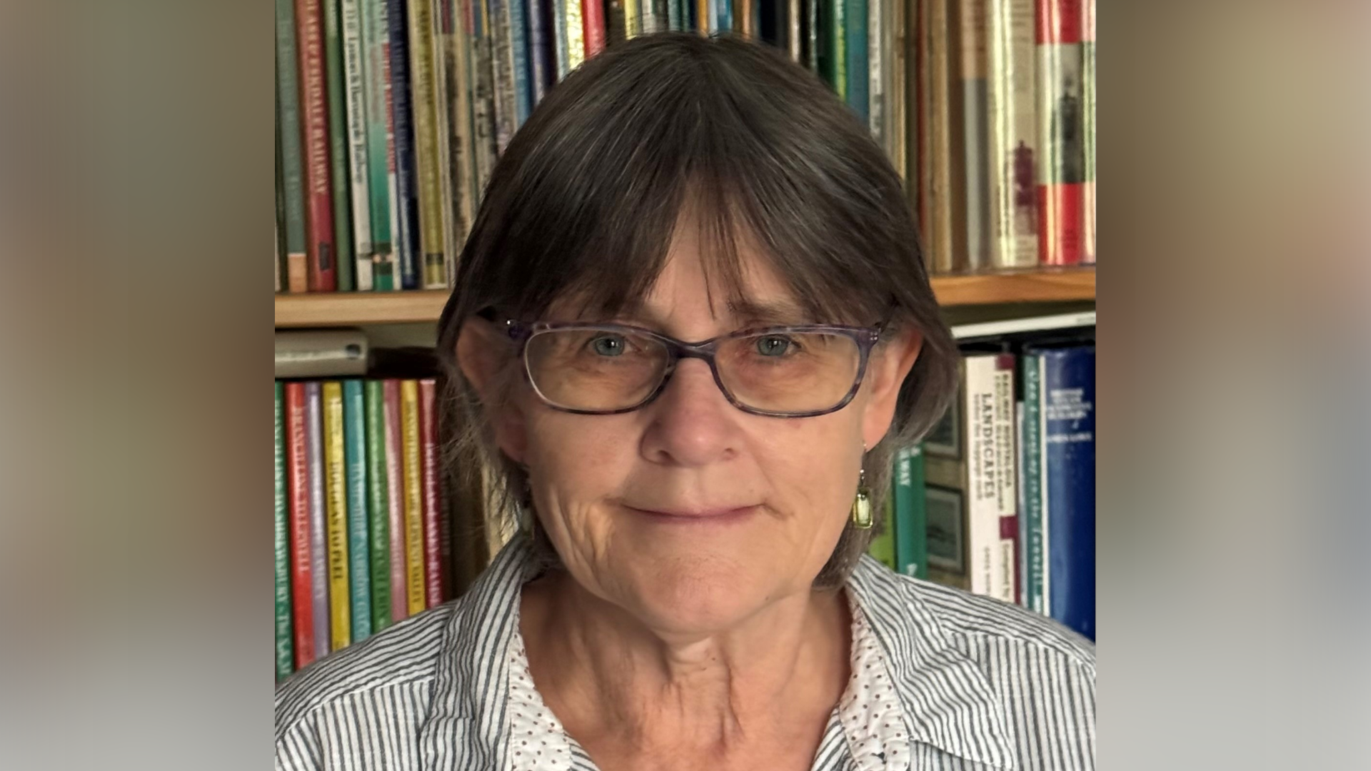Helen Bott smiles at the camera. She stands in front of a bookcase. She has short dark hair and wears glasses as well as a white and blue striped shirt. 