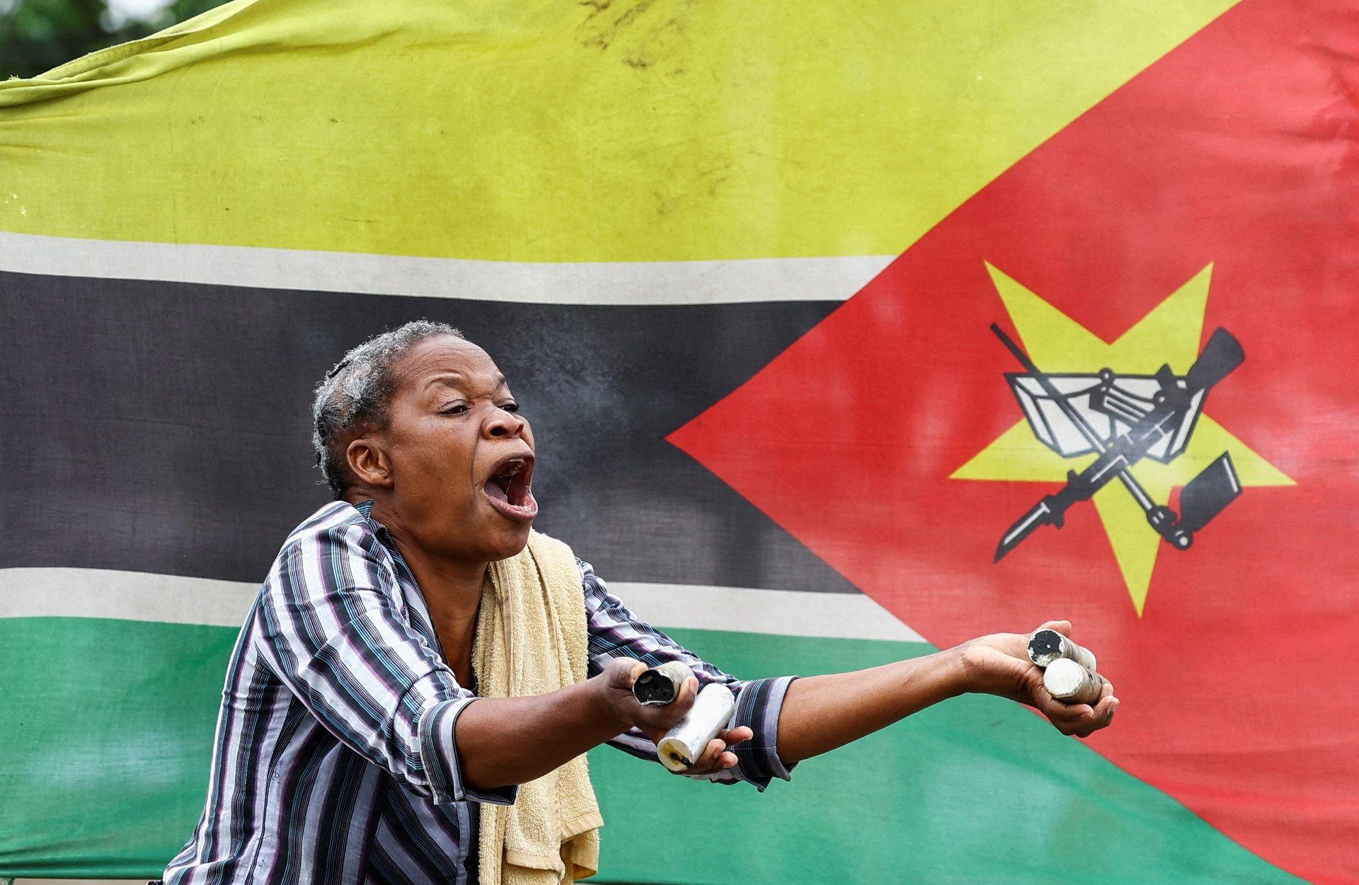 A woman in a stripped shirt with a towel over her shoulder shouts and holds up empty tear gas cannisters at a protest in Maputo. Behind her is the Mozambican flag - Thursday 7 November 2024