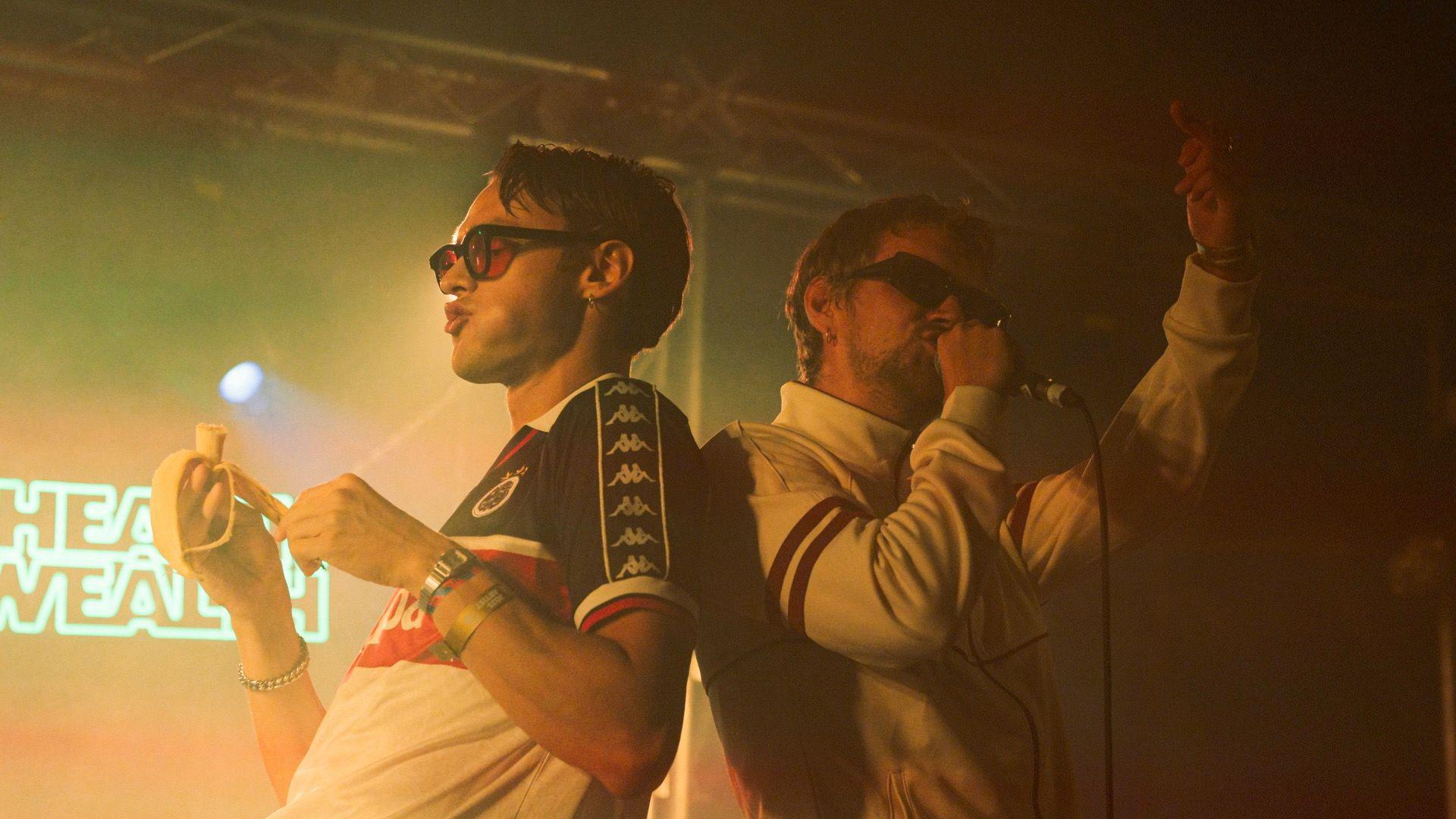 Two male performers are on stage, standing back to back. The back is lit up by a neon sign. The man on the left wears a blue, white and red football t shirt with red tinted sunglasses and is peeling and eating a banana. The man on the right wears a white cotton zip up jacket with red stripes on the slees and black sunglasses. He is holding a microphone up to his mouth, mid-performance, and has one arm raised in the air.