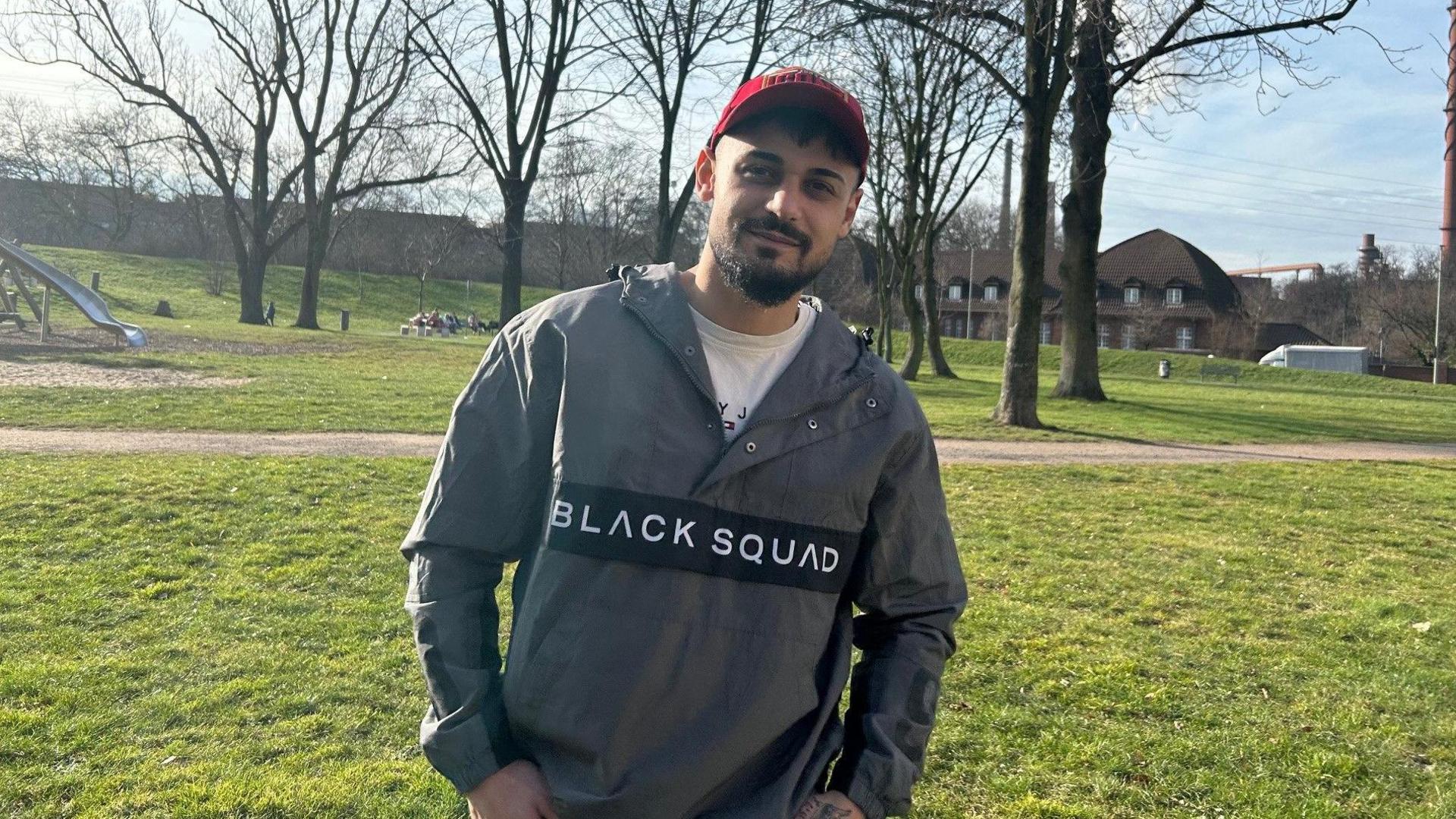 A young man in a red baseball cap stands with his hands in his pockets in a park in western Germany