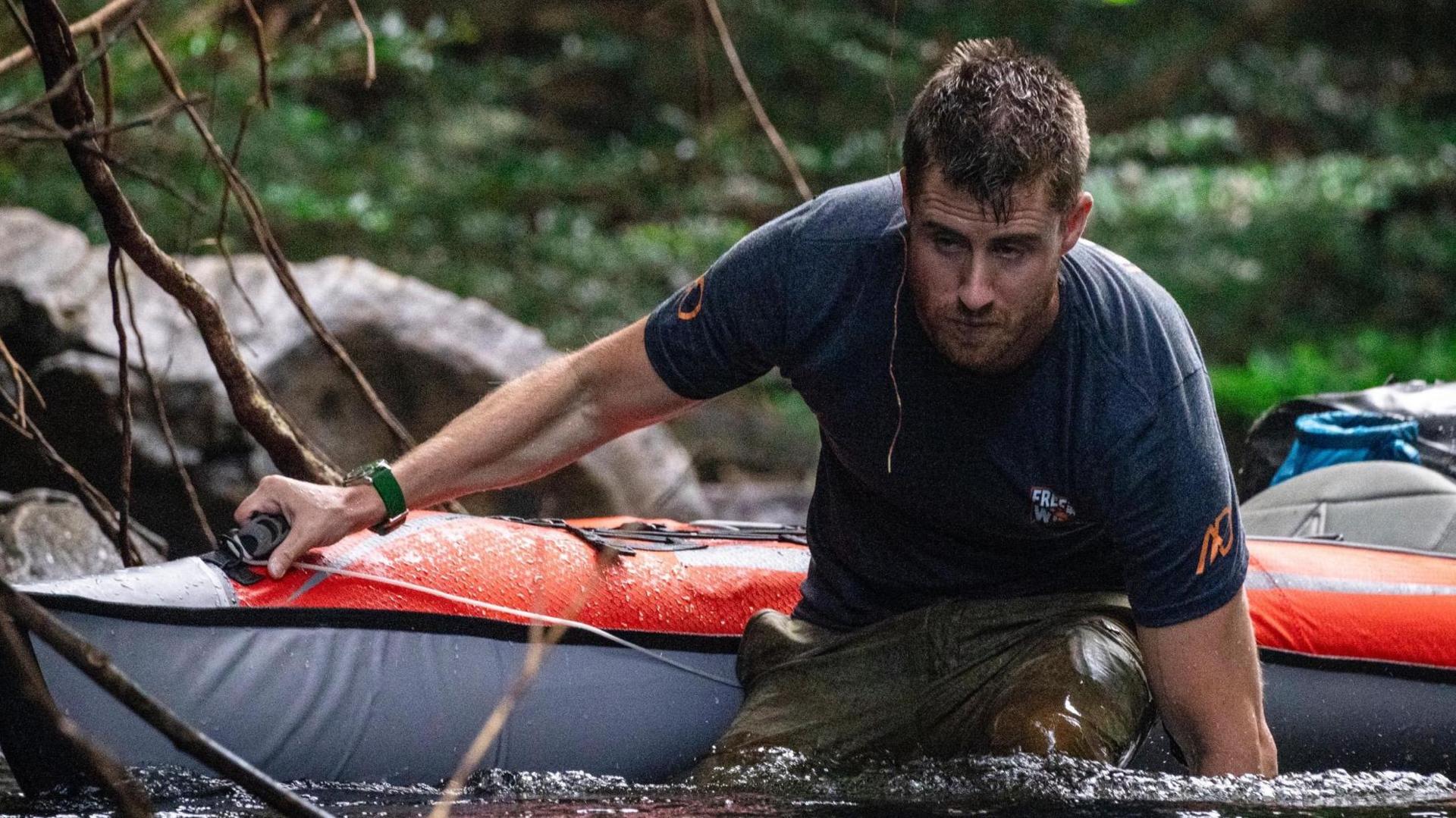 Ash Dykes waist-deep in the Coppename river wearnig trousers and a t-shirt and pulling his kayak with one hand while trying to balance with the other. 