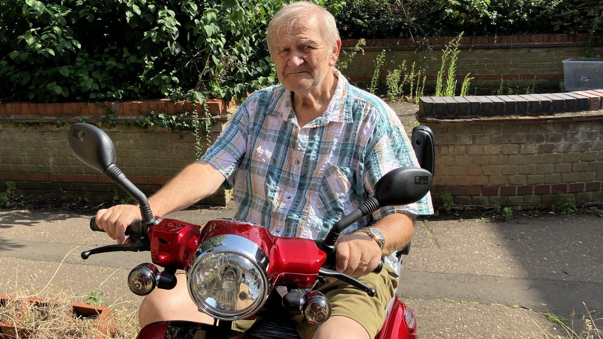 Raymond wearing a checked shirt and brown shirts- sitting on his red mobility scooter 