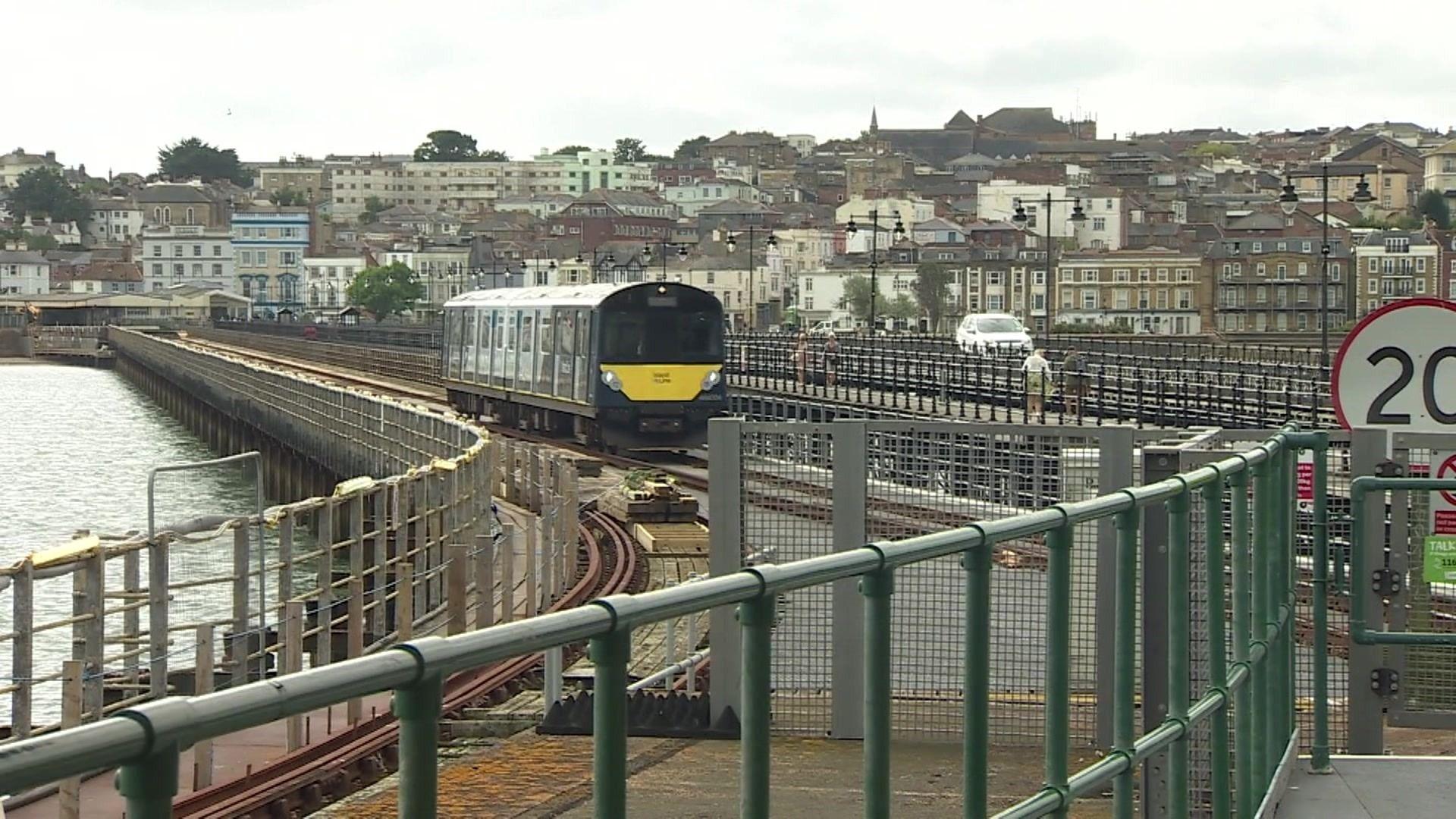 A train goes along a railway line on a pier, with the sea on one side and the buildings on the mainland behind it.