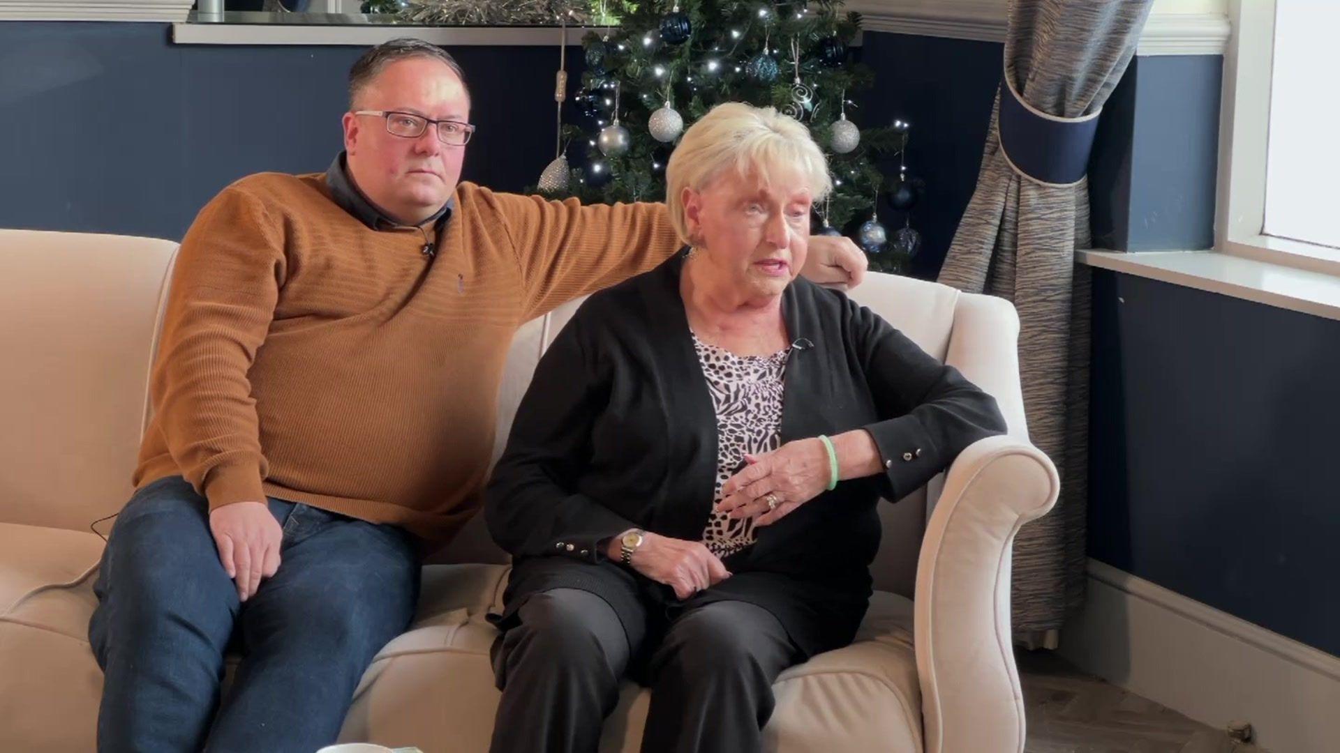 A man in a brown jumper and wearing glasses sits on a sofa beside his grandmother who has blond hair and is dressed in black