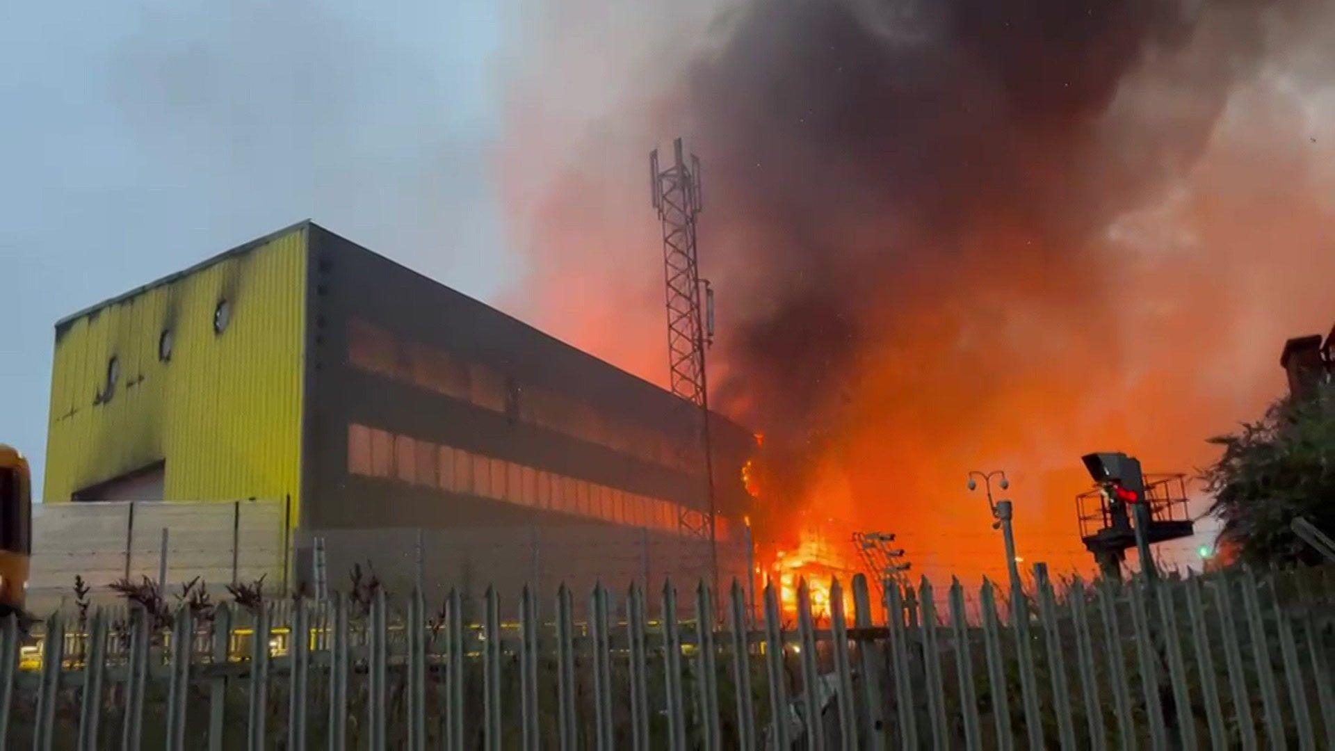 Bright orange flames and thick black smoke engulf a structure behind a metal fence and a telecommunications tower, next to a large yellow and black industrial building under a hazy dusk sky.