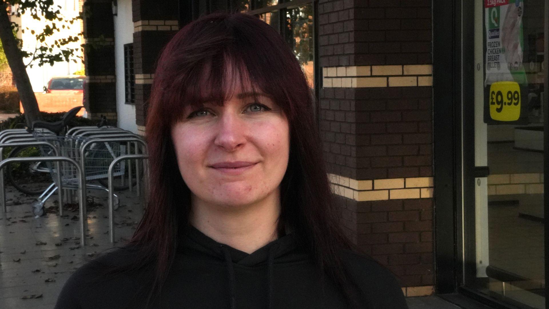Becky Brooker smiles at the camera while standing outside a supermarket. She has long dark red hair with a fringe over her head. She wears a black hoodie.