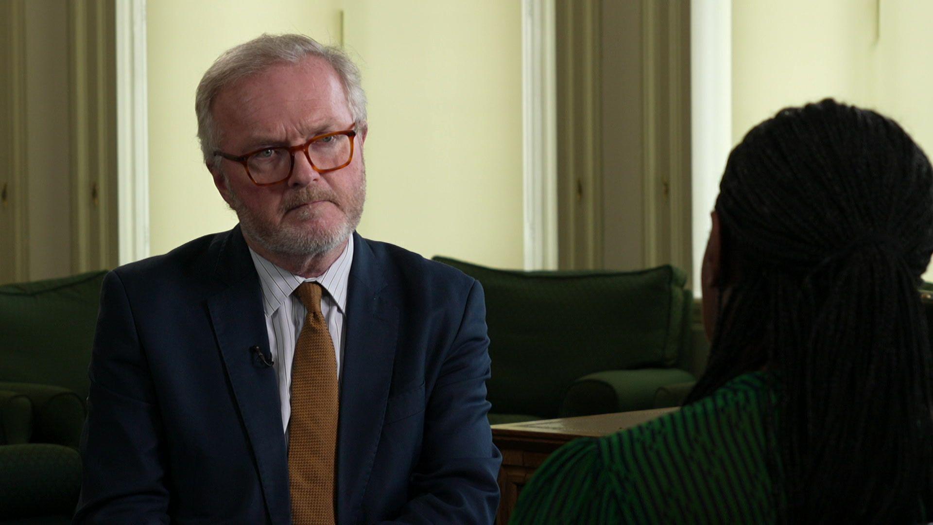 Peter Henley, wearing a dark blazer and gold tie, looks pensively at Kemi Badenoch as she speaks.
