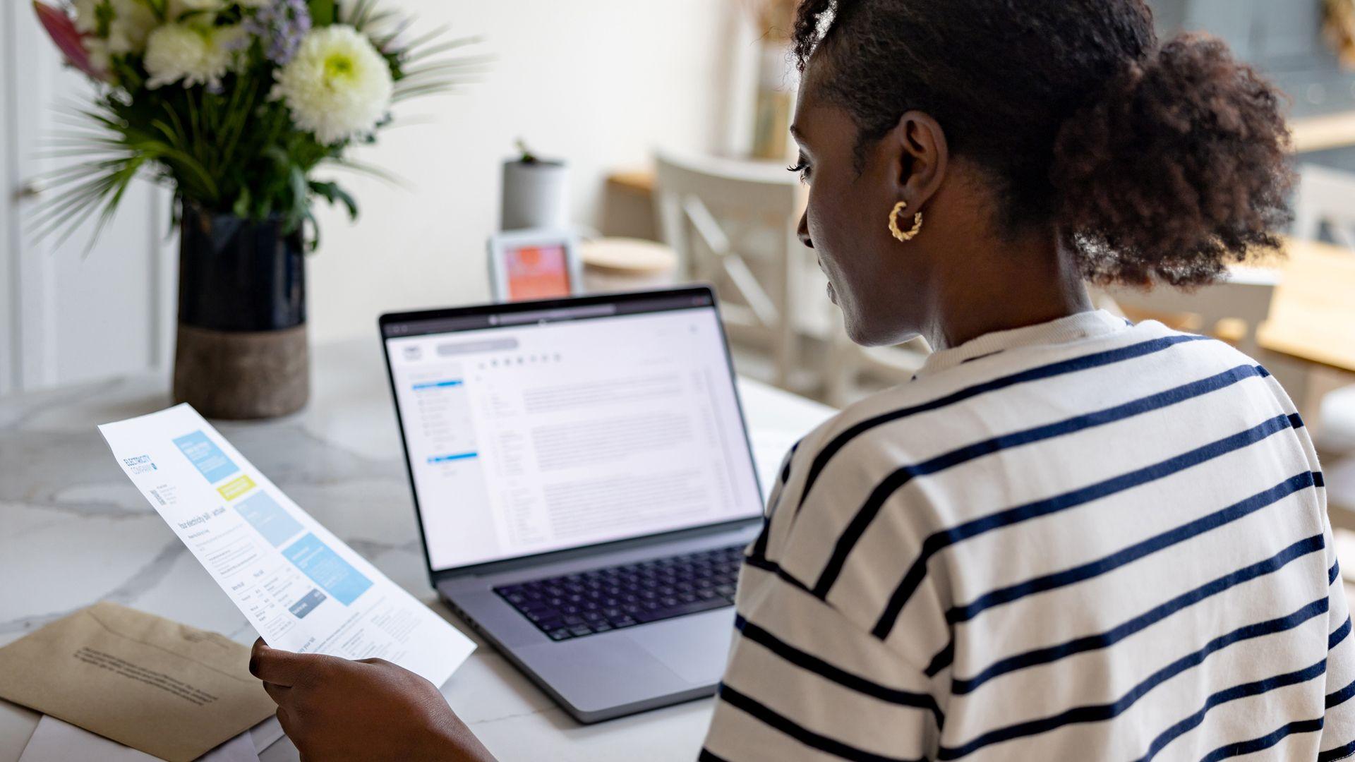 A woman looks at her bills in a letter and on a laptop whilst sat in her kitchen at home
