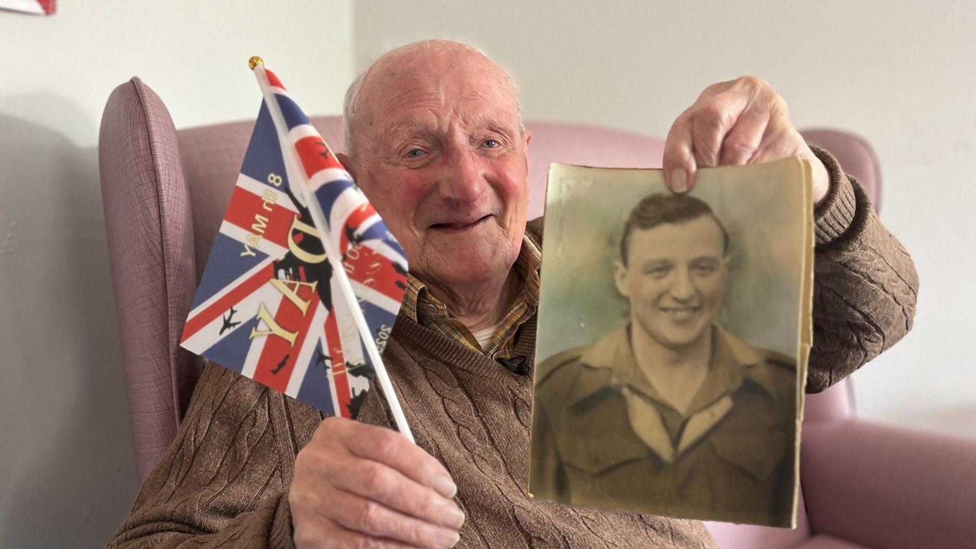 An old, bald, man wearing a brown knit jumper and sat in a pink armchair, smiling at the camera. He is holding up a Union Jack flag and a photograph of him as a young man with short brown hair, wearing a soldier's uniform.