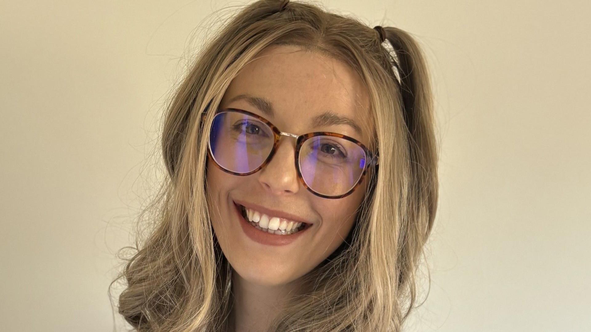 A smiling woman with long blonde hair partially tied up in two bunches. She is wearing black and brown animal print glasses and posing in front of a cream wall. Just her head and shoulders can be seen.