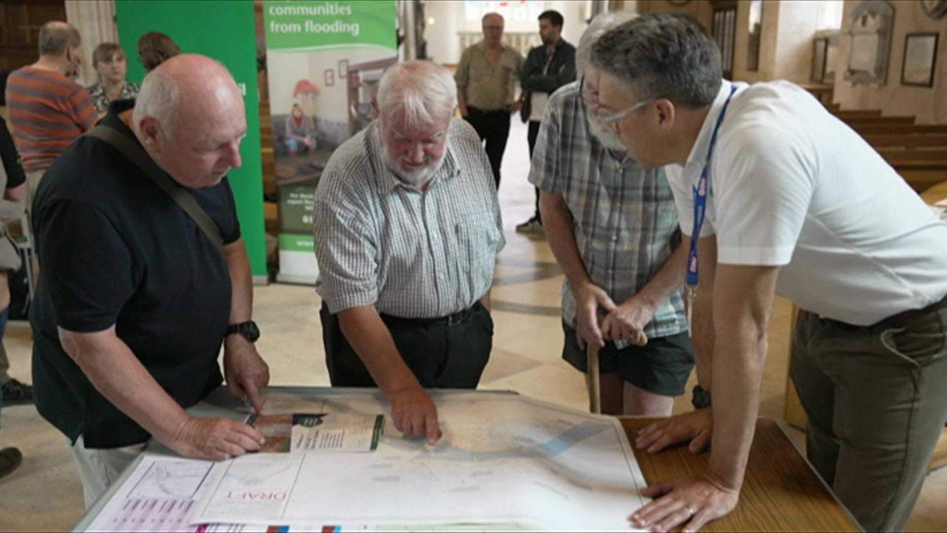 A group of men stand and talk around a table, which has maps and other information laid out in front of them. They are looking and pointing at things.