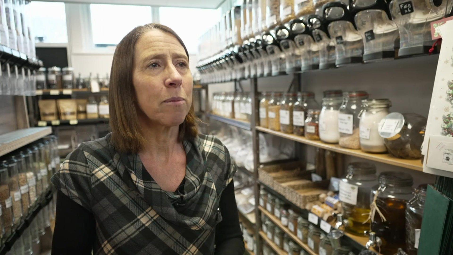 A woman with brown hair looks off to the camera, as she stands in a pantry in a village shop 