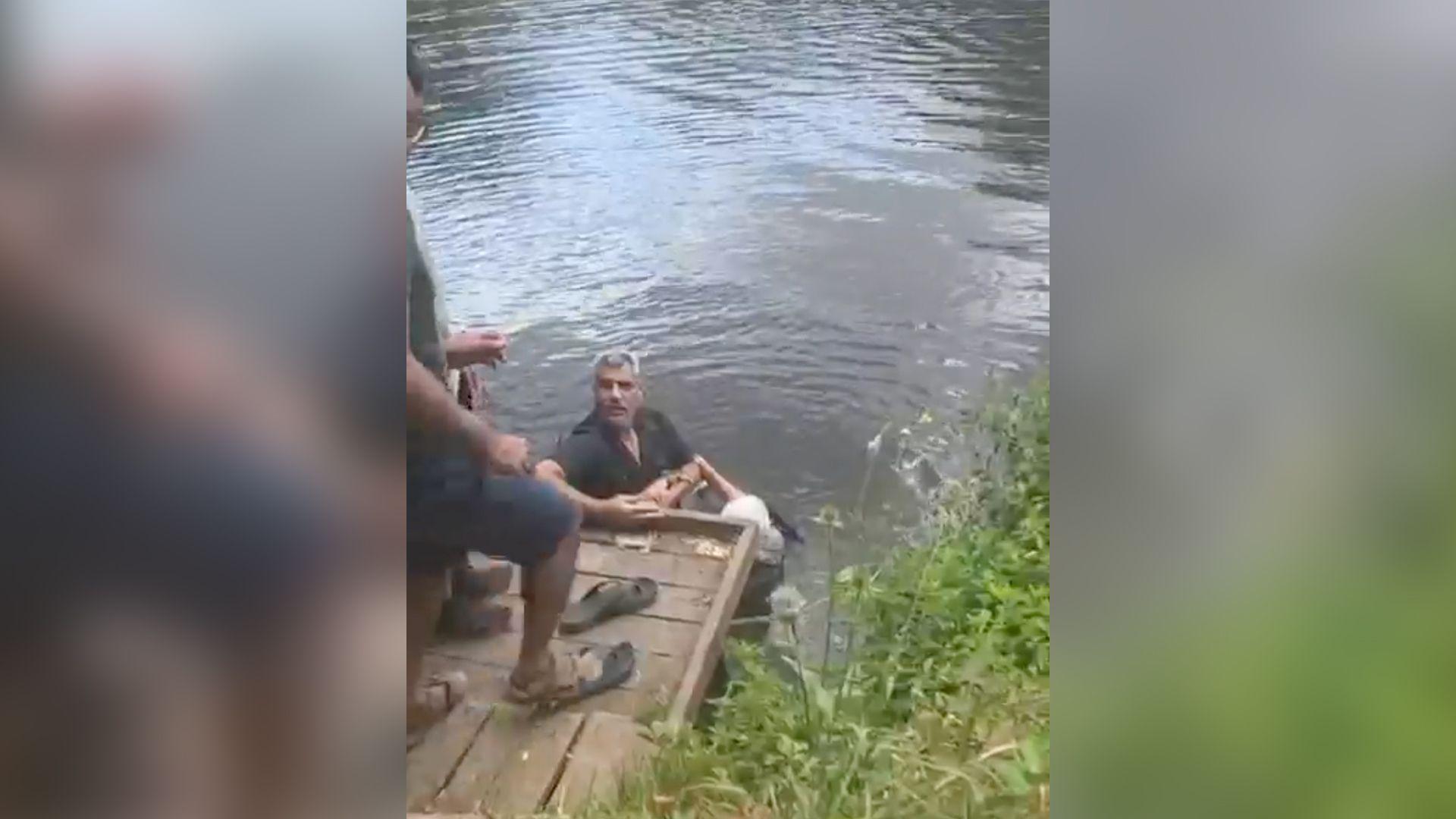 Samer Jouha holding onto an elderly woman in the River Avon in Evesham. He is  holding onto her as he looks up at a short wooden jetty, where other people are coming to assist him.