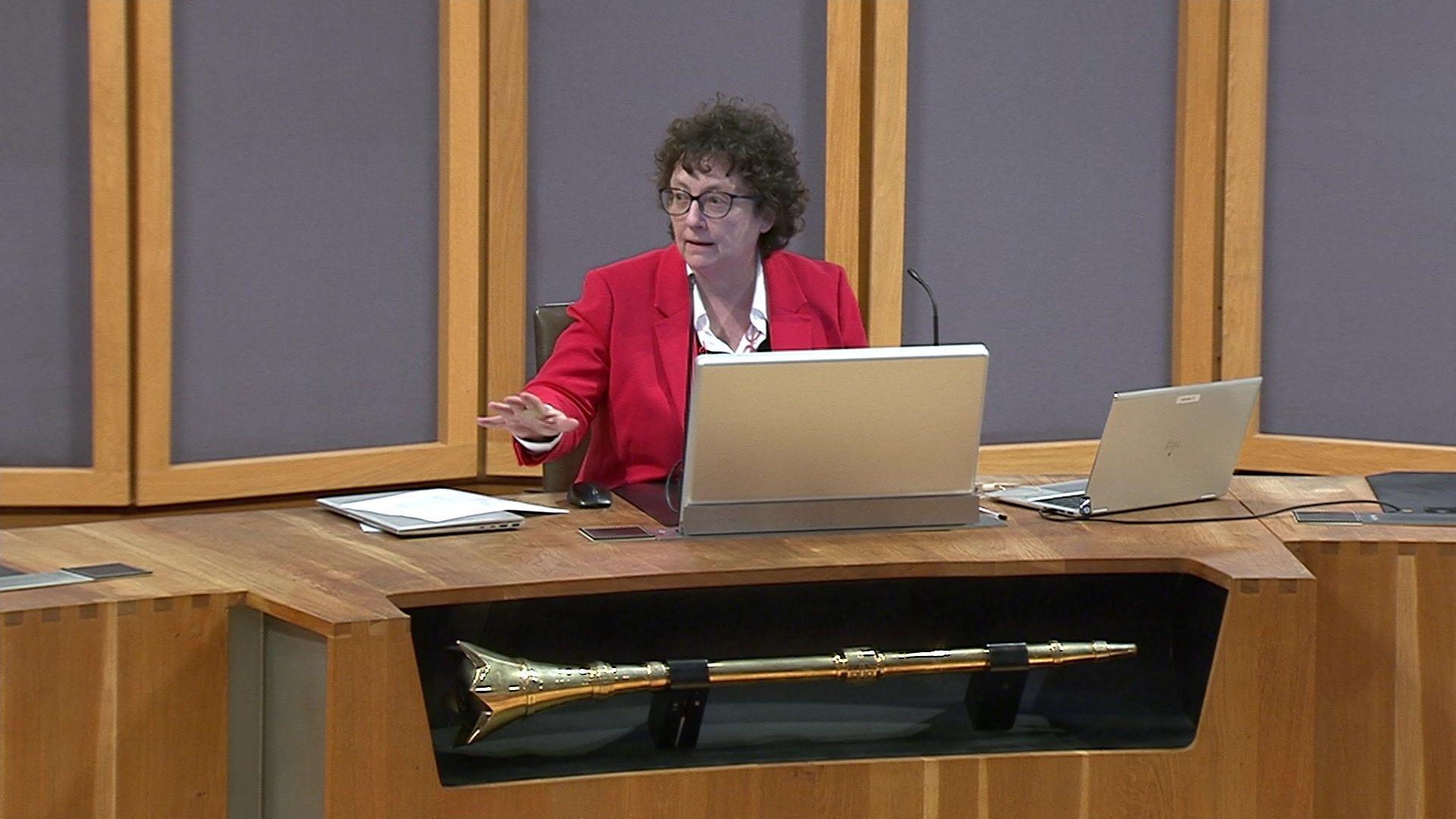 Elin Jones, sitting at her desk chairing First Minister's Questions. She is gesturing with her hand to Darren Millar as she asked him to tone down his words. She is wearing a red jacket, has a computer lap top in front of her and the Senedd's ceremonial mace is in its shelf at the front of the desk