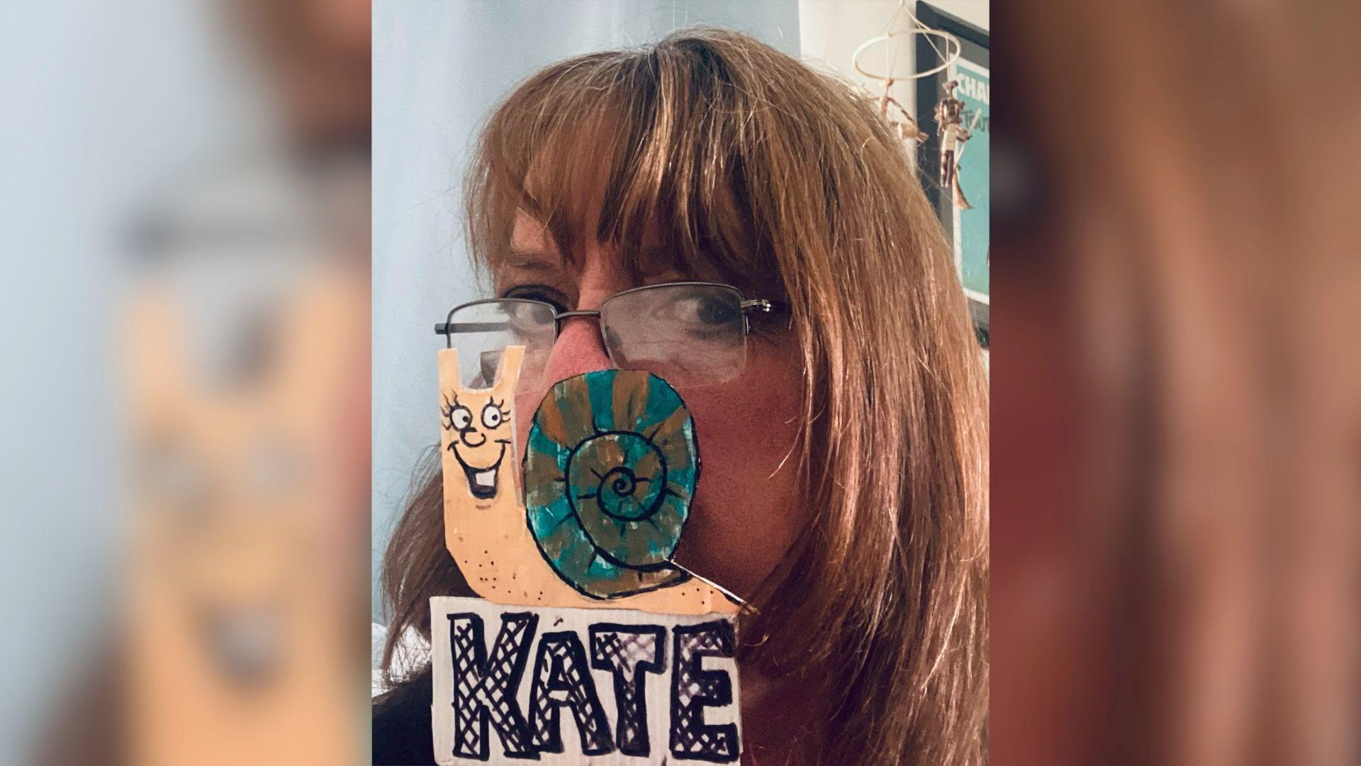 Sue Burleigh holds an illustration of a snail up to her face. She looks at the camera and has rectangular half wire framed glasses on. Sue has brown mid length hair with a fringe. The snail illustration is hiding her nose and mouth. The snail has a cartoon face which is grinning widely showing its teeth and its body is a pale peach colour. The snail's shell has orange and blue stripes and underneath it the name 'KATE' is written in black bubble writing.