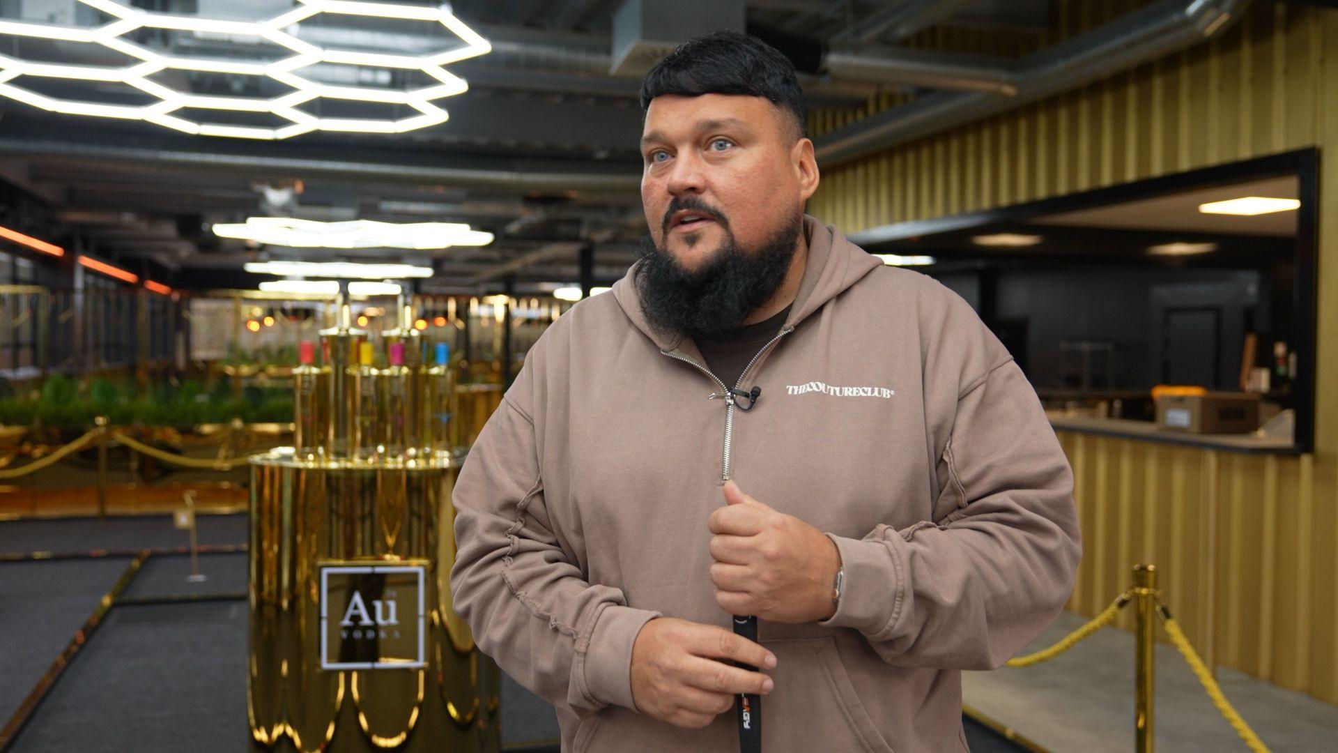 Charlie Sloth in beige tracksuit stands in the Au vodka officers, with gold-wrapped vodka bottles behind him.