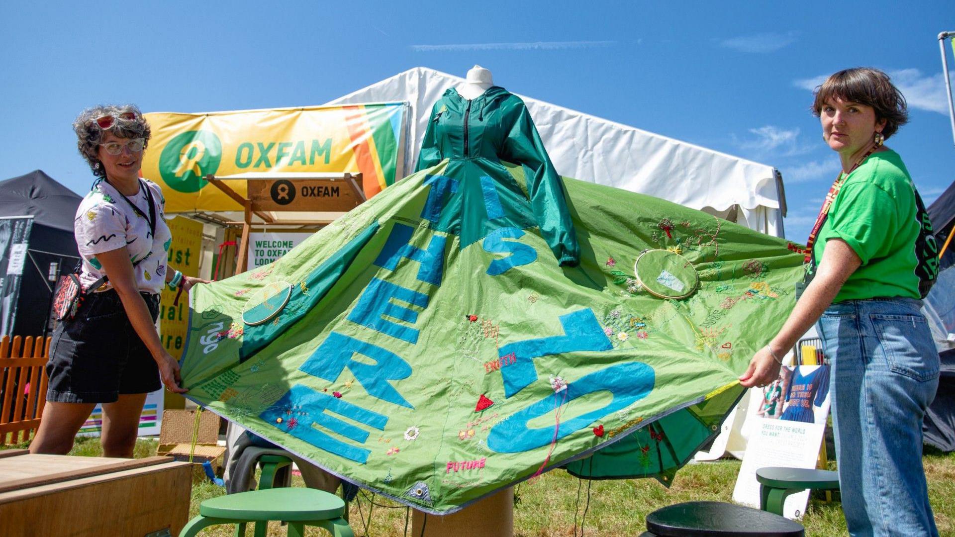 A blue and green dress made out of tents with messages on is showcased by designer Alice Bowen-Churchill outside of an Oxfam tent on a sunny day.