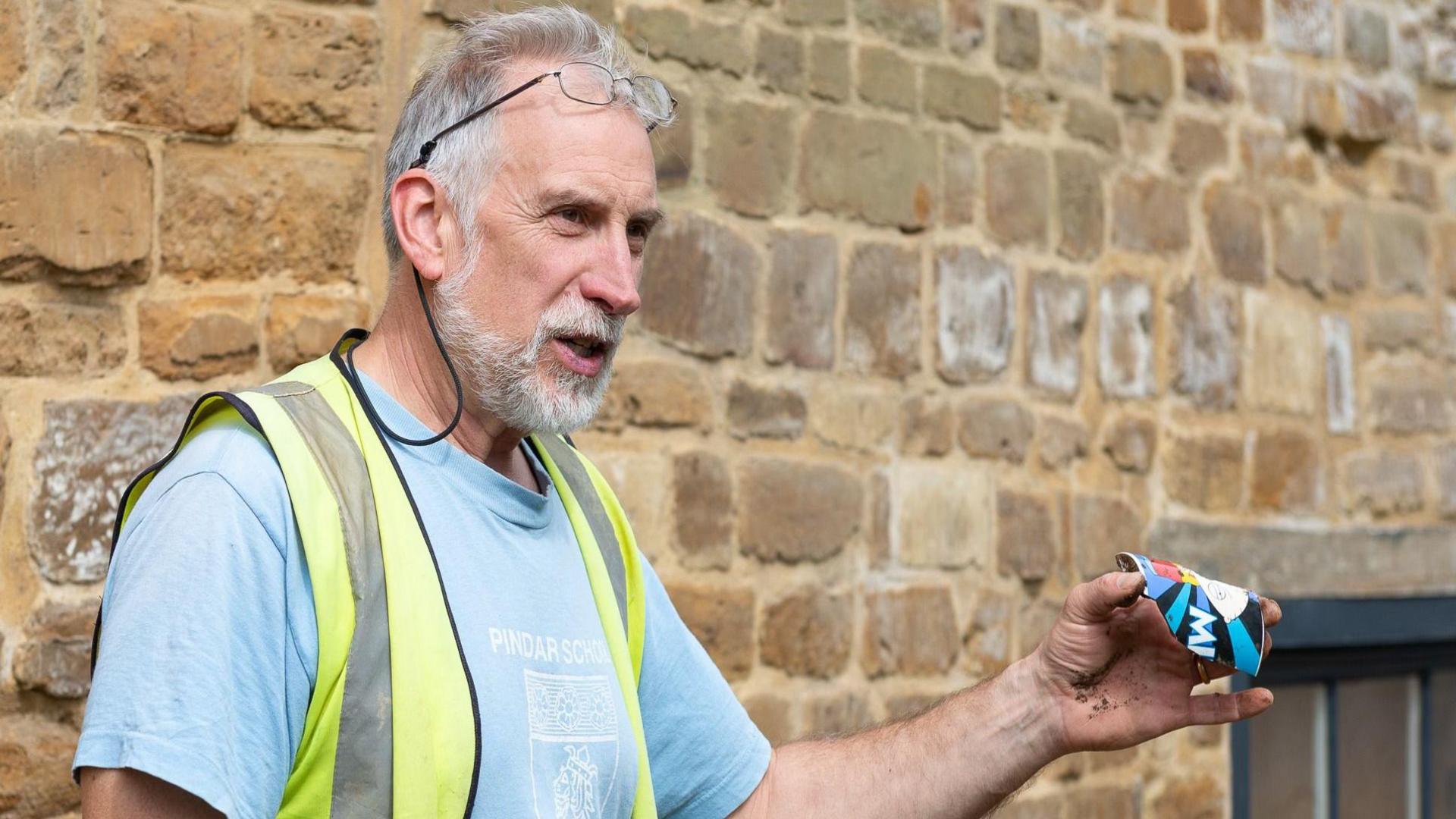 John Buglass with short white hair and beard wearing a yellow hi-vis and holding a crisp packet