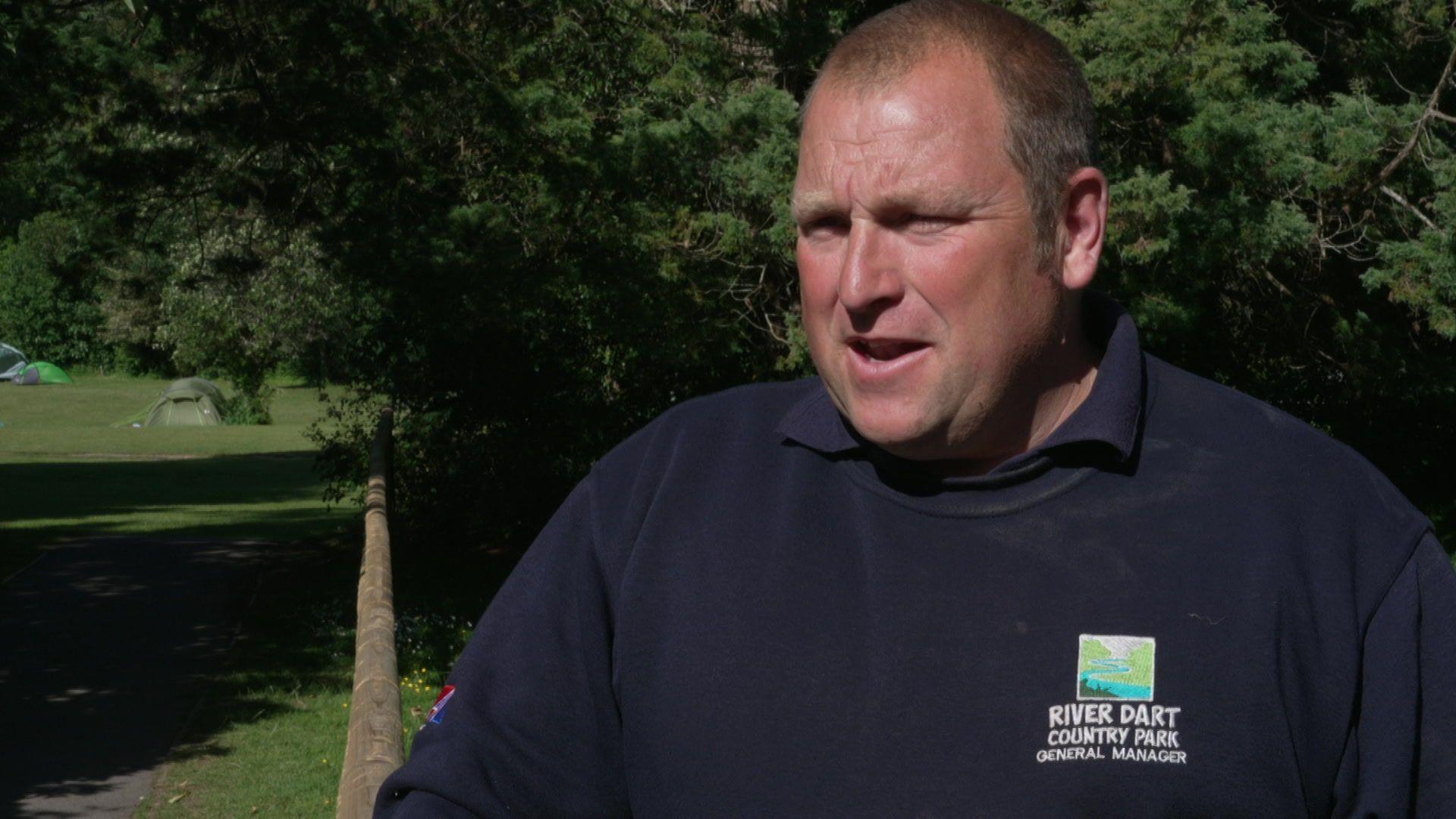 A man in a blue top stands next to a wooden fence and trees behind.