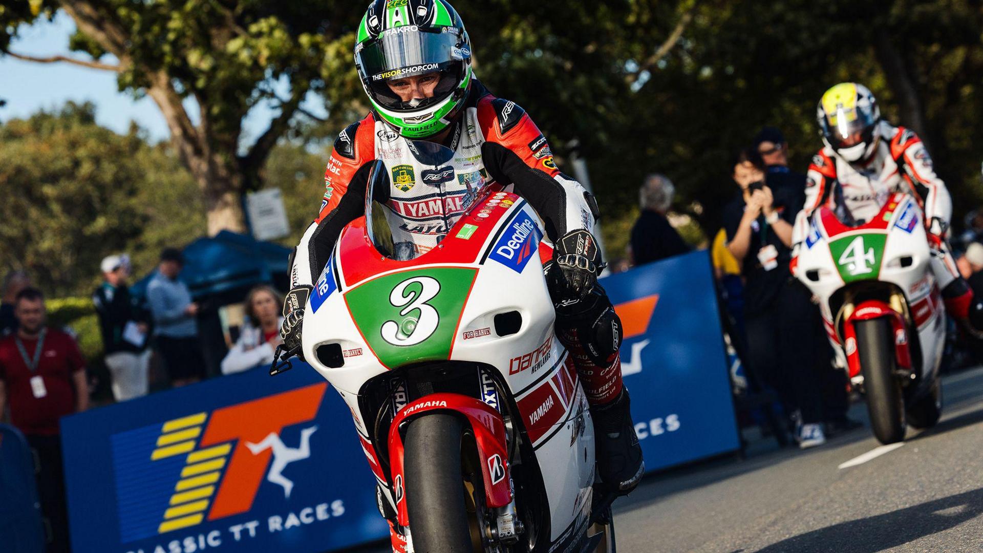 A rider in colourful leathers and a helmet rides on a motorcycle, another follows behind on a tarmacked road. Fans look on from behind a blue barrier which reads Classic TT Races in in white writing, with a blue, yellow and red logo.
