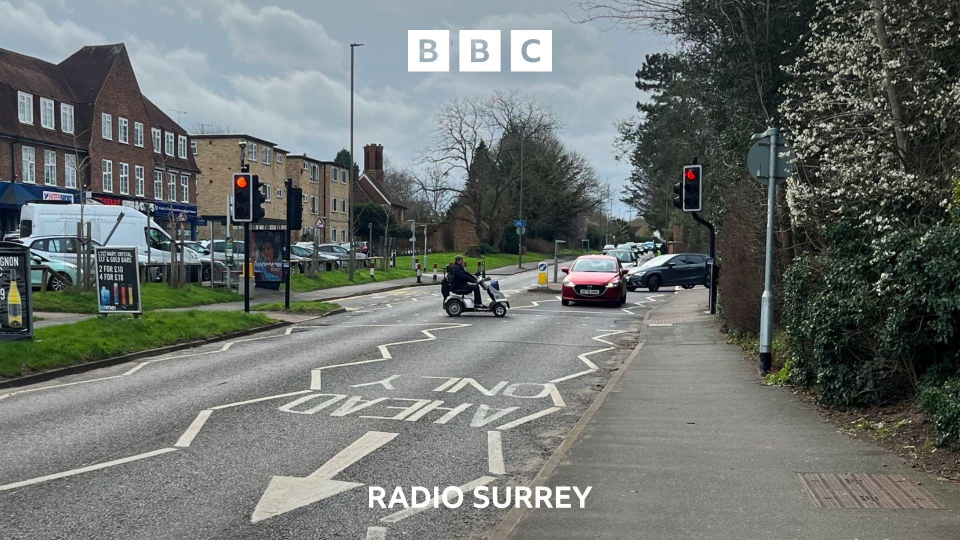 A person on a mobility scooter crosses a single carriageway road at a pedestrian crossing. A red car stops to wait at the traffic lights. There are shops in the background.