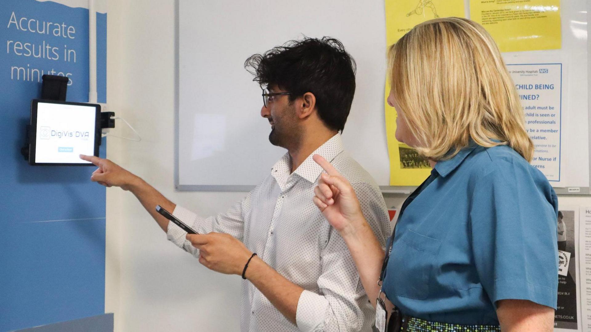 Jamie Mistry is standing with Louise Allen in front of an eye test screen in a hospital. He has dark hair and a beard and is wearing a light-coloured shirt. He is holding a mobile phone in his left hand. Louise Allen is standing behind him. She has fair hair and is wearing a blue blouse