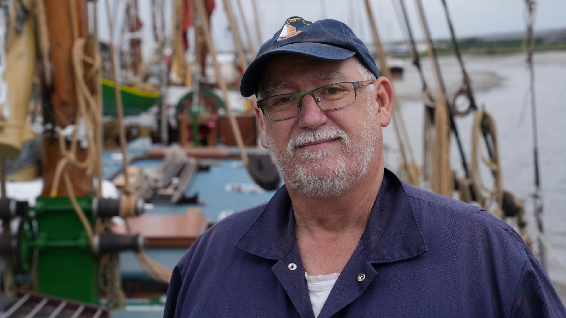 Tim Kenney is dressed in a navy sailor's jacket, wearing a navy baseball cap, and standing on the deck of Pudge the Thames sailing barge