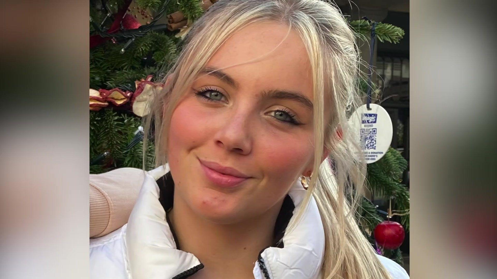 A head and shoulders shot of a young woman with blonde hair tied back in a pony tail who is smiling at the camera. She is wearing a white coat and standing in front of an Christmas tree.