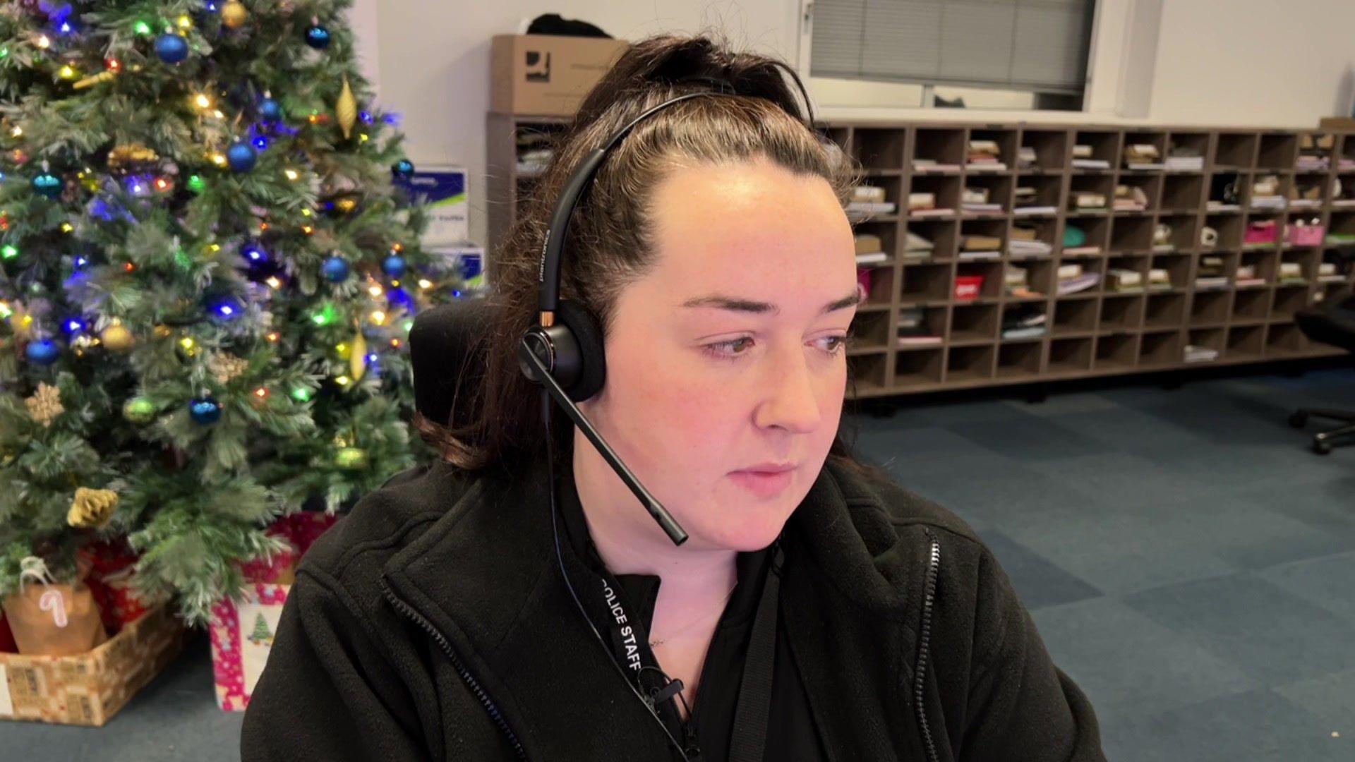 A woman sits in a call centre wearing a headset with a Christmas tree behind her