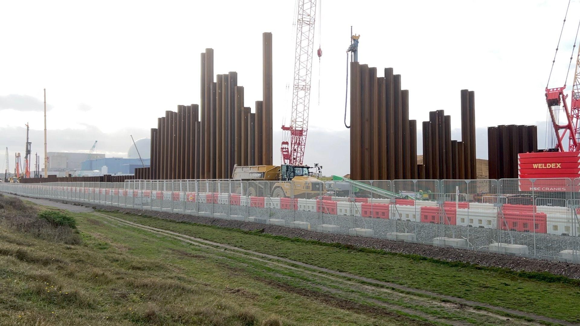 The construction site of Sizewell C, with Sizewell B and A in the background. There is a tipper truck and cranes on site, with large metal piles going into the ground.