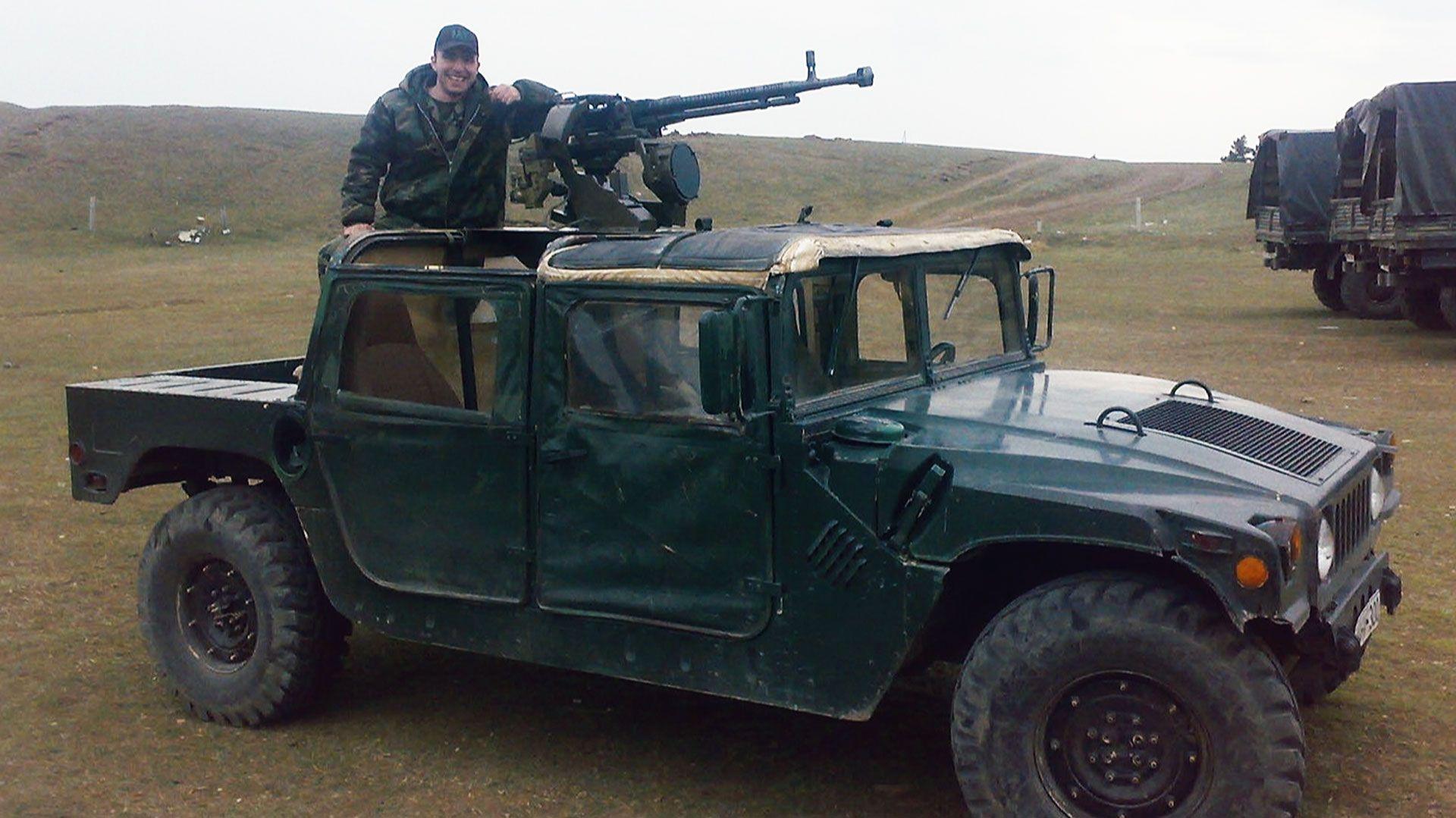 Lasha wearing army fatigues and a cap standing on an armoured vehicle and resting his arm on a mounted rifle